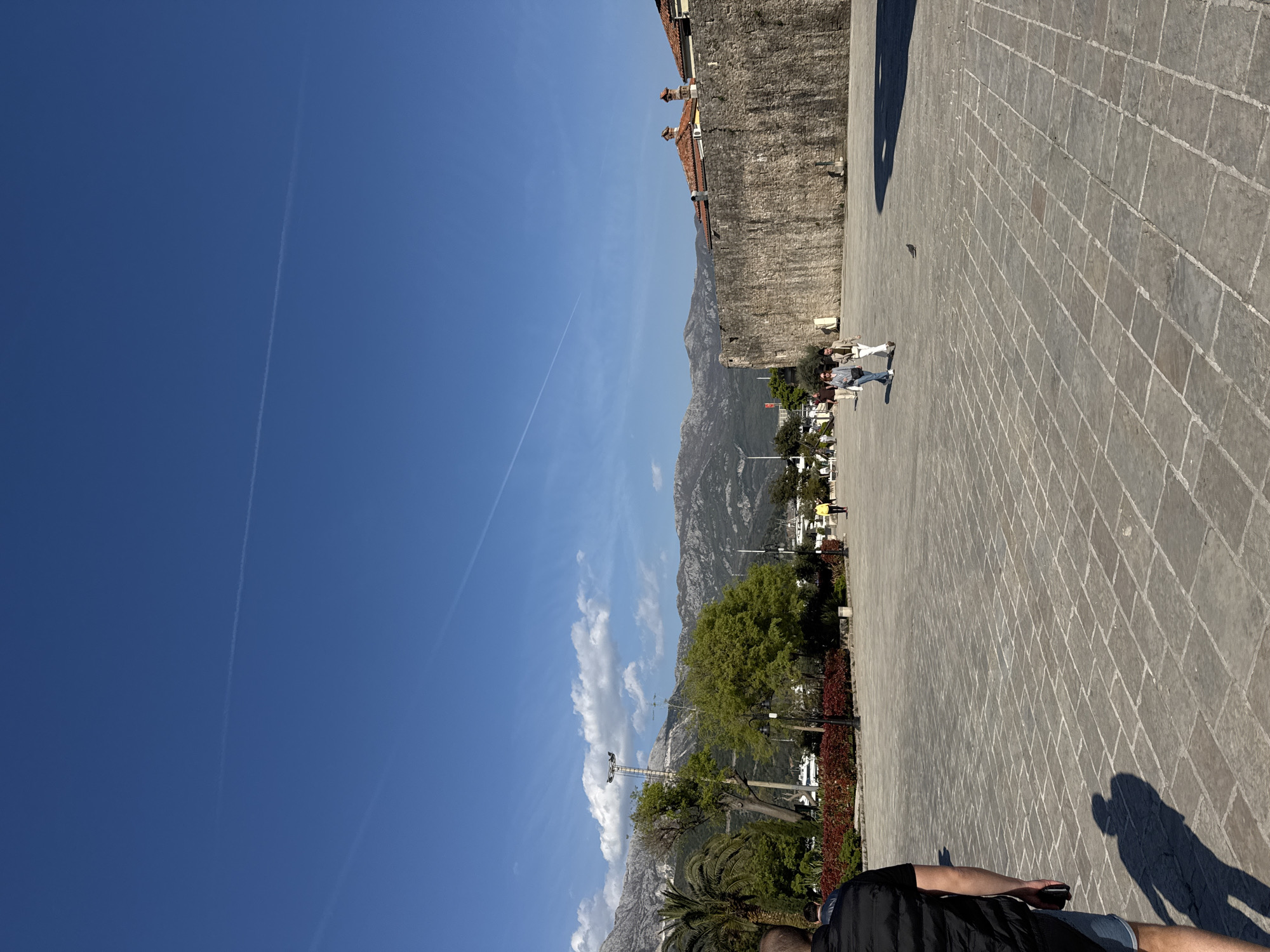A photograph shows a public square in the city of Budva, Montenegro. The sky is clear blue with contrails. In the background, there are mountains. The paved square is on the right side of the frame. Several people are walking on the square. Buildings with brick-red roofs are visible near a stone wall on the right. Trees and vegetation separate the square from the mountains in the distance. An individual is standing in the foreground on the right.