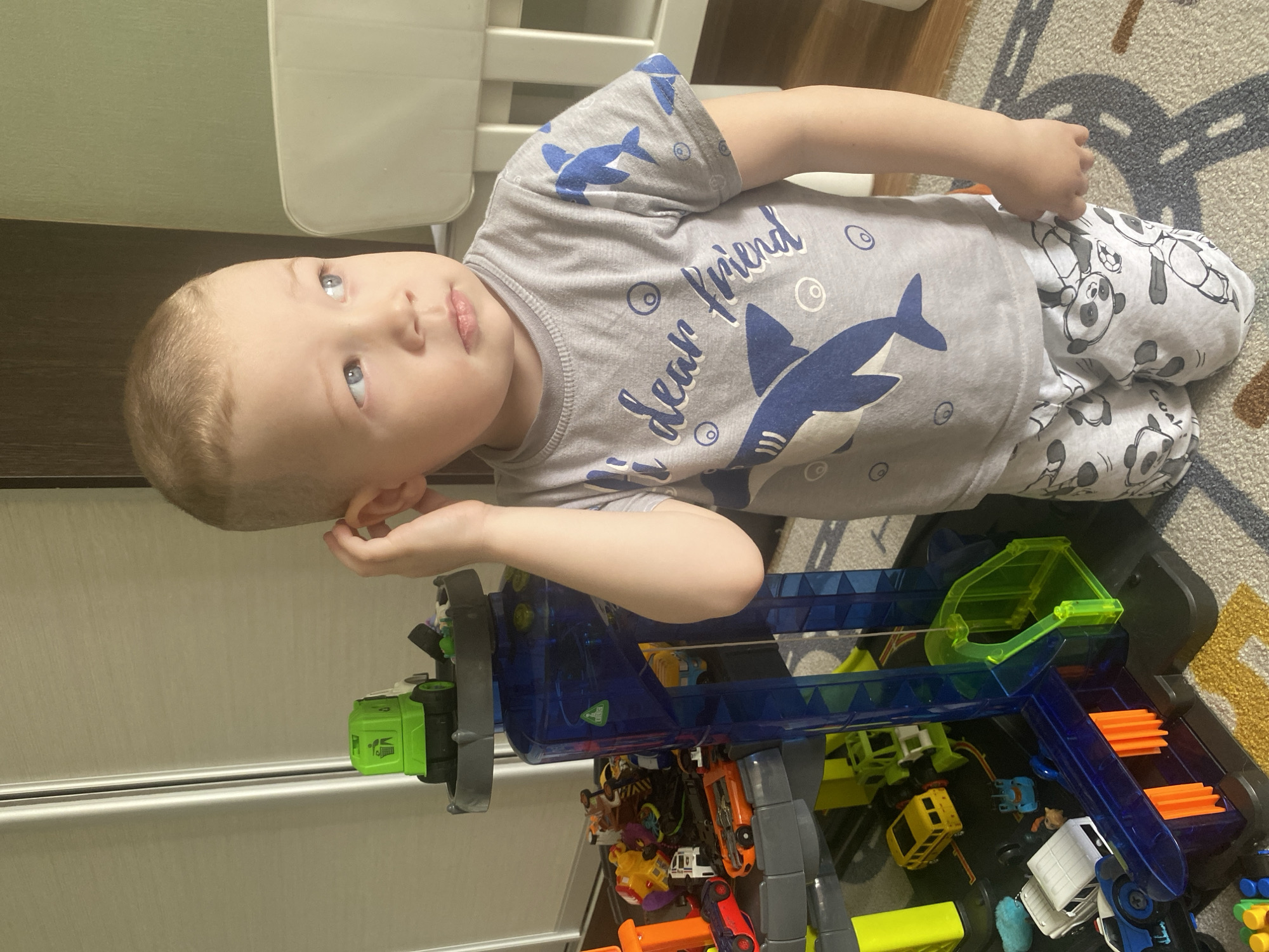 A young boy lies on a carpeted floor next to a toy car track and collection of toy cars. The boy, fair-skinned with light brown hair and blue eyes, wears a gray t-shirt with a shark design and panda-print pajama pants. He rests his left hand near his ear and looks upward. The toy car track is primarily blue and green, with multiple levels and assorted toy cars beneath. A carpet with a gray and white pattern is visible under the boy and track. The location is not discernable as Sochi, Russia, from the visual elements of this image.