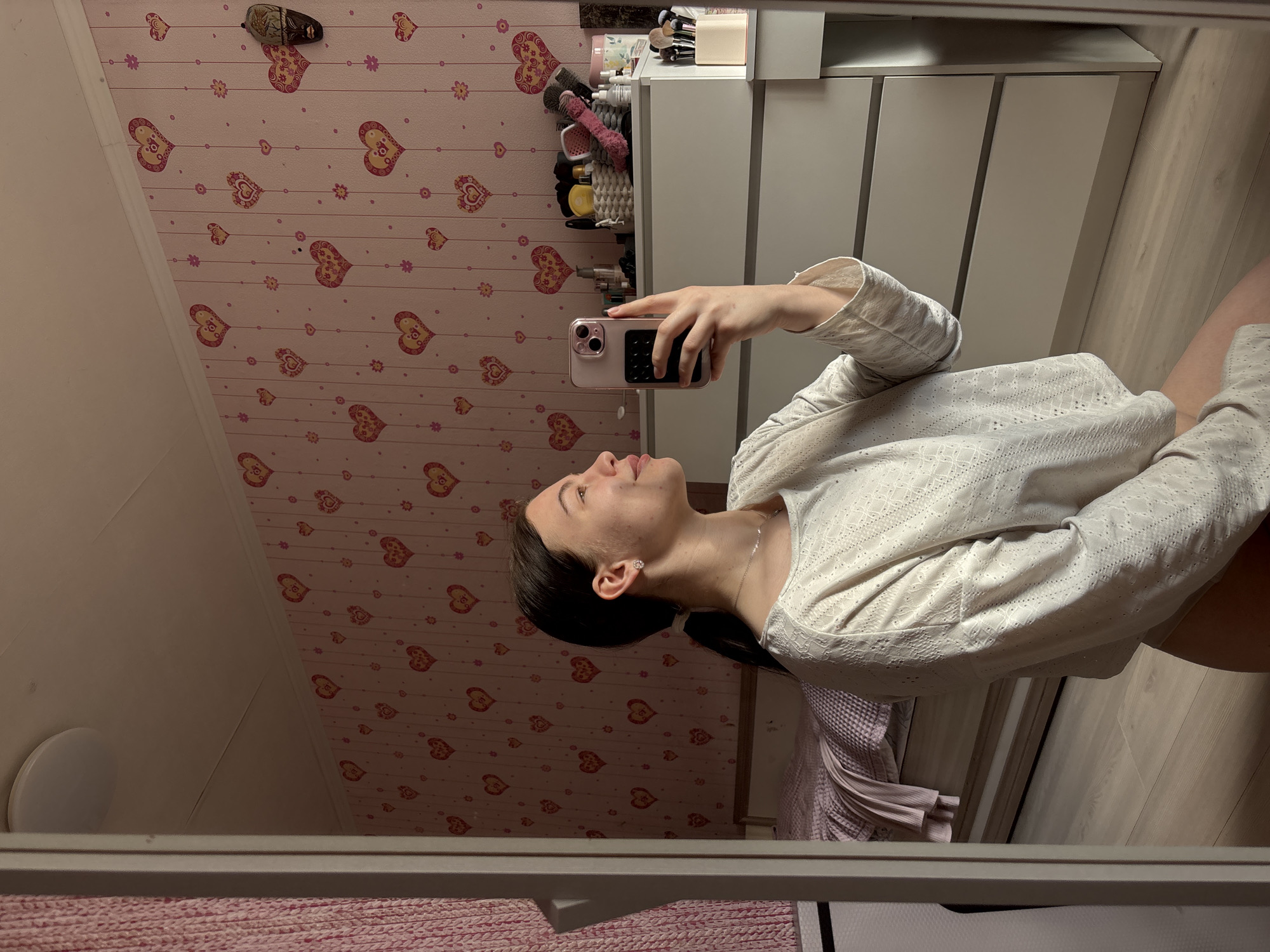 A young woman is taking a selfie in a mirror. She is holding a pink iPhone with a black camera grid. She is wearing a white, textured long-sleeve shirt. Her hair is dark and pulled back in a ponytail. She is wearing a silver necklace and small earrings. The background consists of a pink wallpaper with hearts, a white dresser with toiletries on top, and wooden floors. The mirror’s frame is visible at the bottom. The location appears to be a bedroom in a house in Perm, Russia.