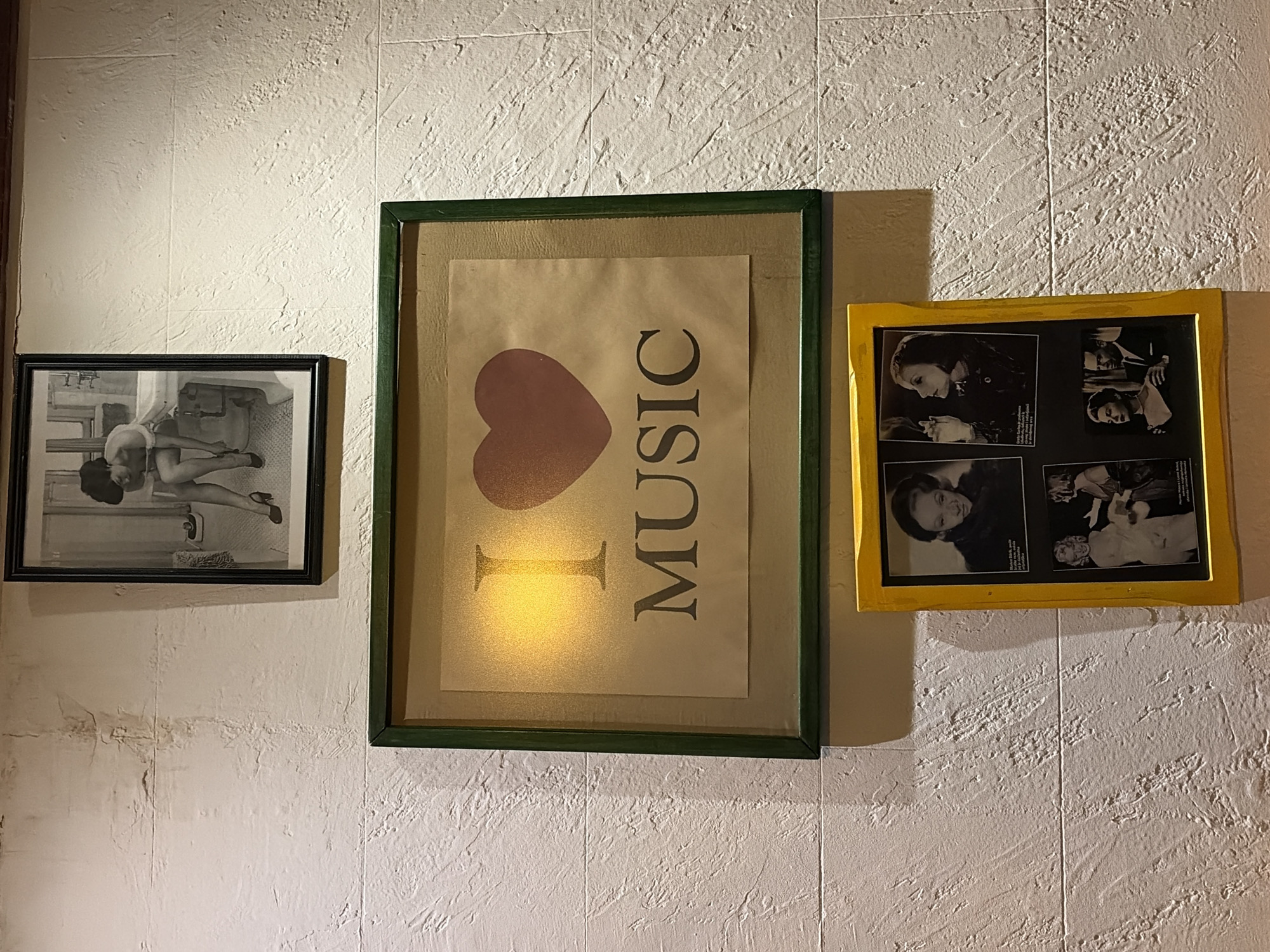 Three framed artworks hang on a textured, off-white wall. From left to right: a black-and-white photograph of two people in a bathroom setting; a framed tan-colored poster with a red heart and the word "MUSIC" in dark brown, within a green frame; and a yellow-framed collage of black-and-white photographs featuring people's faces. The location is likely a building interior in the city of Bar, Montenegro, as determined by the prompt. The scene is a wall decoration consisting of various framed artworks.
