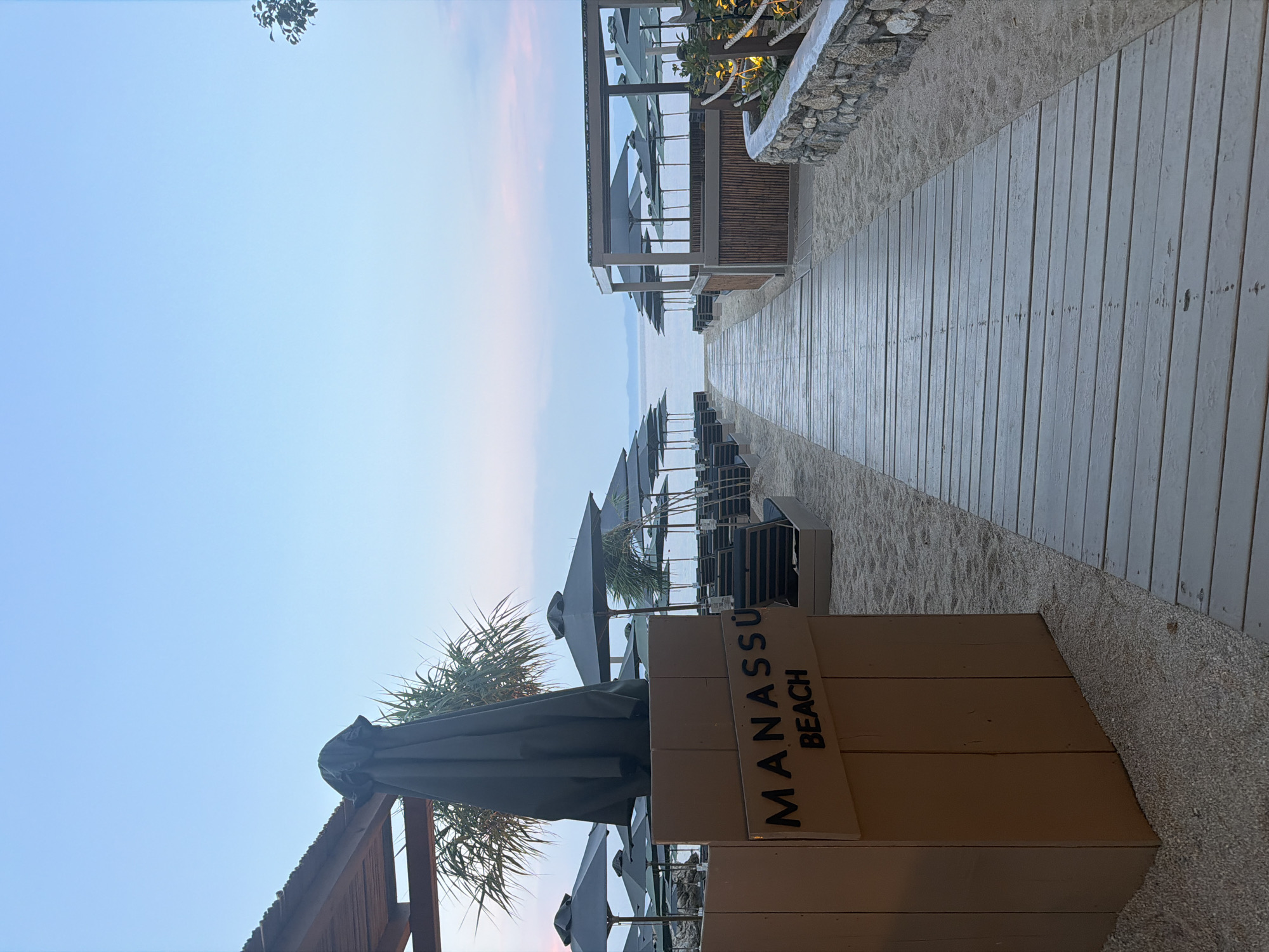 The image shows a tranquil beach scene in Sithonia, Greece. A wooden walkway extends towards the sea, lined with rows of closed beach umbrellas and lounge chairs on either side, suggesting a quiet, perhaps early morning or late evening atmosphere. On the left, a wooden signpost reads "MANASSÜ BEACH" in black lettering. Behind the sign, a large, dark umbrella is folded and covered. The beach is composed of fine sand and small pebbles. In the background, the calm sea meets a pale blue sky with hints of pink and purple hues near the horizon. A structure resembling a beach bar or canopy with thatched walls and umbrellas is visible on the right. The overall impression is one of serene beach resort ambiance.
