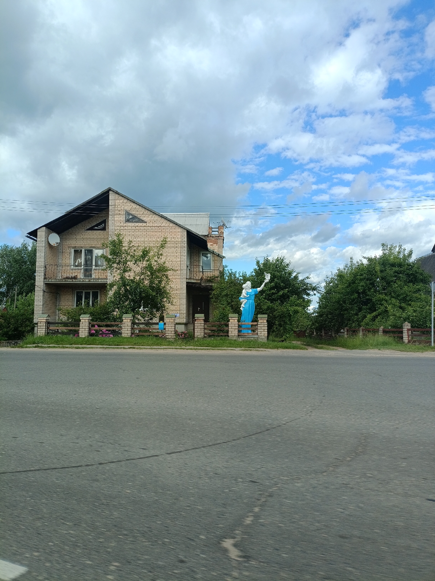 A two-story brick house stands on the left side of the image, with a satellite dish on the upper left corner and a balcony on the second floor. The house has a dark-colored roof and several windows. A statue of a woman in a blue dress is positioned to the right of the house, holding a bird or object in her hand. Both the house and the statue are surrounded by green trees and bushes, and a wooden fence separates the property from the road in the foreground. The road appears to be asphalt and shows some wear and tear.
The sky is partly cloudy, with a mix of bright blue patches and large, white, and gray clouds. The lighting suggests it is daytime, likely late morning or afternoon. There is no visible text in the image. The scene appears to be a residential area in a rural or suburban setting. The overall mood is calm and serene.