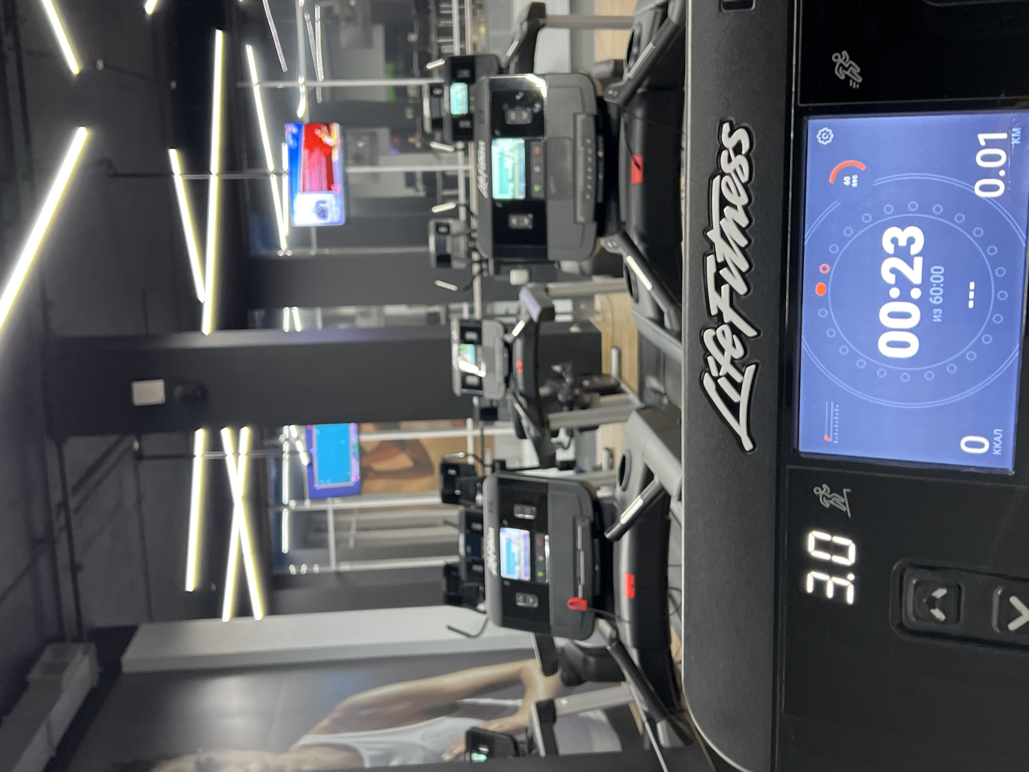 The image captures a modern gym or fitness center, specifically a row of treadmills. The perspective is from the side of one treadmill, looking down the line of machines. The room features dark walls and ceiling, illuminated by striking linear fluorescent lights arranged in geometric patterns.
In the foreground, a black Life Fitness treadmill console is prominent, displaying digital metrics. The screen shows "00:23" (likely elapsed time), "из 60:00" (out of 60 minutes, indicating a timer or session length), "0.01 KM" (distance), and "0 ККАЛ" (calories, zero in this instance). A speed or incline setting of "3.0" is visible. An icon of a running person and a settings cog are also present on the display.
Further down the line, several other treadmills of the same model are visible, each with its own screen, some appearing to show video content with prominent red and blue colors. Reflections on the floor or a large poster show parts of a person's body, possibly indicating exercise or promotional imagery. The overall impression is one of a clean, well-lit fitness facility. Given the text "из 60:00" and "ККАЛ", the location is consistent with the provided context of Khimki, Russia.