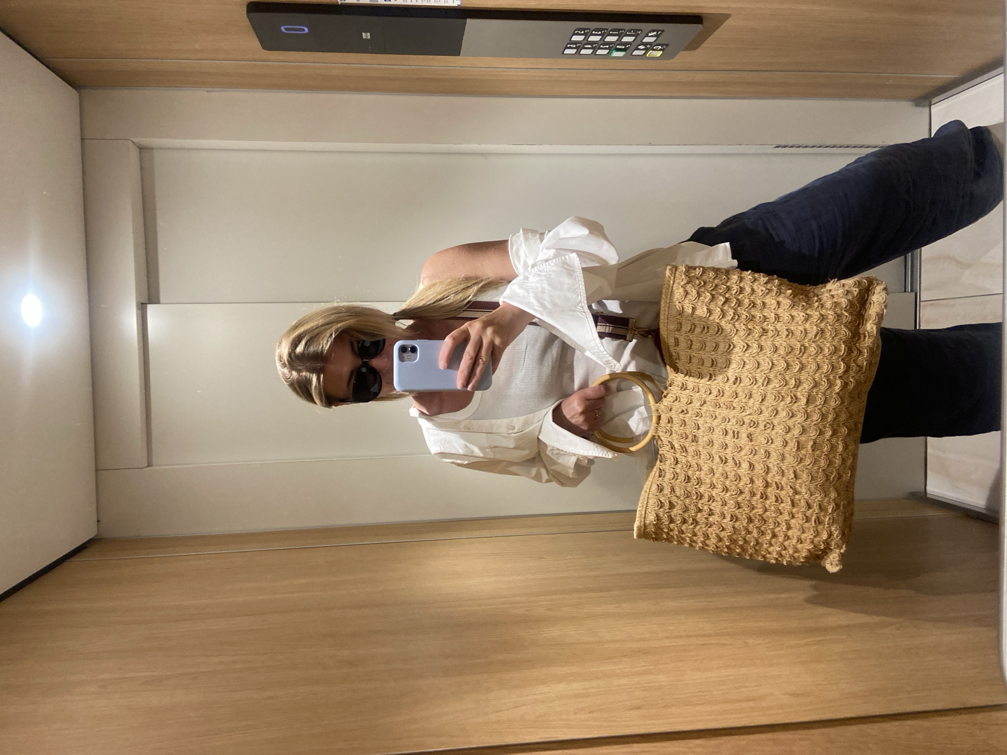 A woman takes a mirror selfie in what appears to be an elevator. She is wearing sunglasses, a white shirt, and dark-colored pants. She is holding a light blue phone with her right hand, and a tan woven bag with a circular wooden handle hangs from her left arm. The elevator has a wooden floor and light-colored walls. Above the mirror is an elevator control panel. Although the image is not geotagged with a location, the request mentions the city of Budva, Montenegro. It is likely that the woman is in an elevator somewhere in Budva, Montenegro.