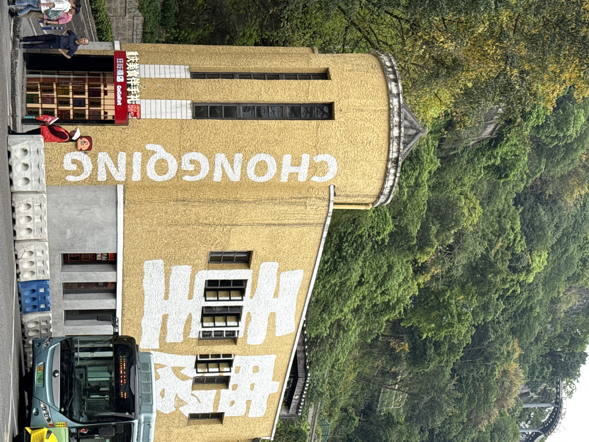 The image captures an urban scene in Yuzhong District, China, featuring a prominent yellow building, a bus, and lush green hills. The large, textured yellow building, possibly part of a tower or a uniquely shaped structure, occupies the left and central parts of the frame. It is adorned with large white painted text. Vertically, the Chinese characters "重庆好礼" (Chongqing Good Gifts) are visible, and horizontally, the English word "CHONGQING" is displayed. Multiple rectangular windows are interspersed across the building's facade.
A red illuminated sign is mounted above an entrance on the building, displaying Chinese text "重庆美食伴手礼 找吃商店 GoGoEat" (Chongqing Delicacies Souvenir Shop GoGoEat), with "CHONGQING LOCAL GIFTS" written below in English. Near the building's base, a person in a red jacket and hat is visible.
In the foreground, a blue and gray public bus is partially seen on the street, with its front displaying illuminated symbols. Its license plate, "渝A 17790F," is clearly visible, indicating its registration in Chongqing. Grey, patterned concrete barriers line the street next to the bus.
Several people are visible in the upper left of the image, standing or walking on an elevated walkway or street, observing the area.
The background is dominated by a densely vegetated hillside, covered in a vibrant array of green trees and foliage, suggesting a rich natural landscape surrounding the urban development. Hints of a bridge or elevated structure can be seen through the trees on the right side. The scene is set during the daytime, under natural light, depicting a lively integration of urban life with the natural environment.