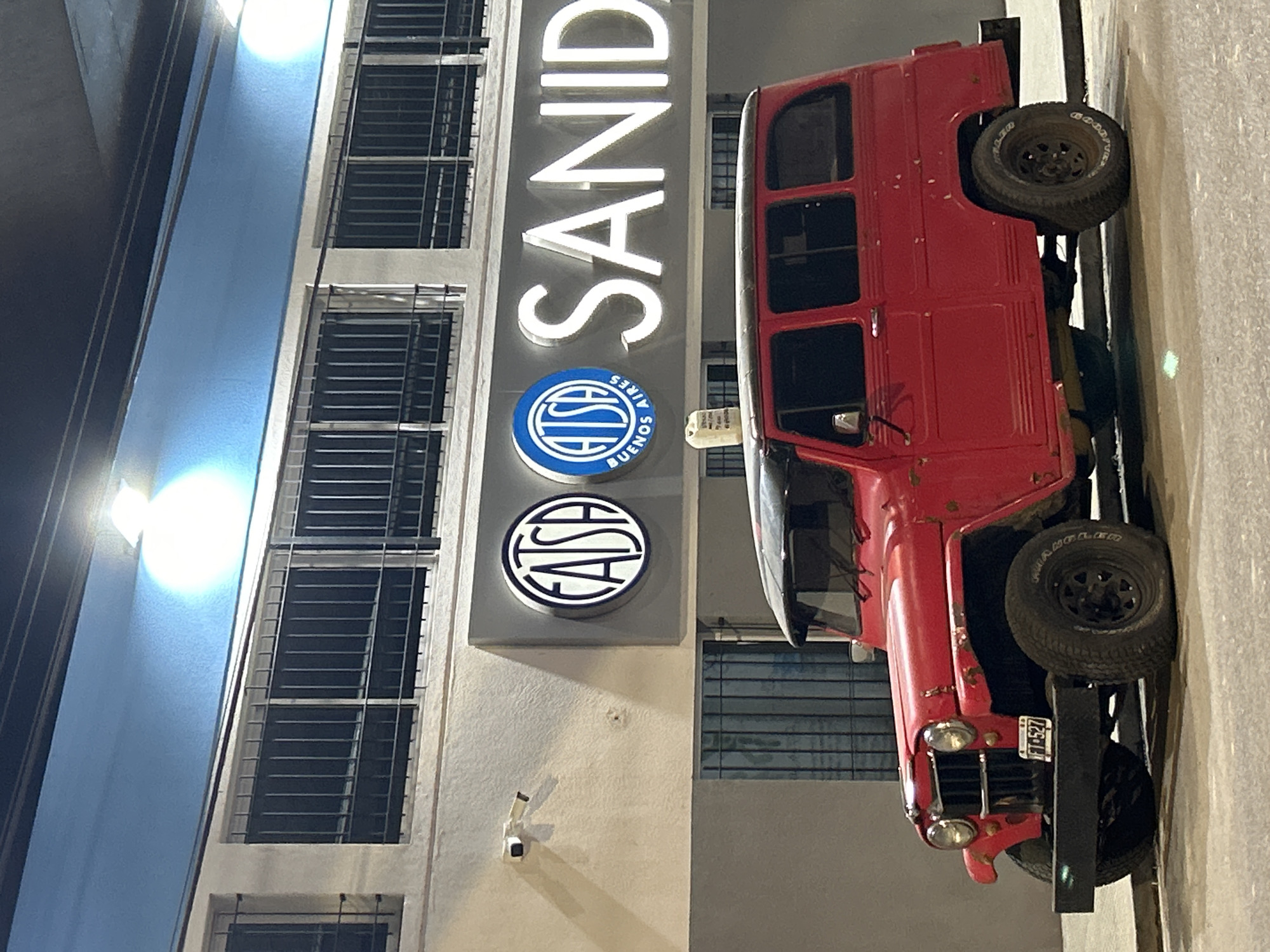 The image shows a vintage red station wagon parked in front of a building with illuminated signs. The station wagon has a black bumper and black wheels. The building has a light blue facade on the upper portion and white on the lower. Above the vehicle, a neon sign displays "FATSA" in a circular logo and the word "SANIDAD" in large, illuminated letters. Another circular logo adjacent to "FATSA" appears to be "ATSA BUENOS AIRES." A security camera is visible on the exterior wall of the building. The scene is outdoors, at street level, under artificial lighting, suggesting it is nighttime or dusk. The license plate of the car reads "FTG-527".