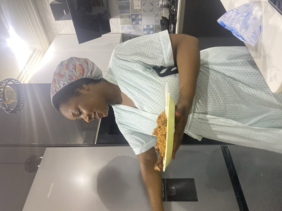 The image, rotated 90 degrees clockwise, presents a woman in what appears to be a kitchen or dining area. She is focused on a disposable foam plate in her hand, which holds a serving of a reddish-brown rice dish, likely jollof, accompanied by a piece of chicken. The woman is wearing a light-colored, short-sleeved robe or house dress with a subtle dotted pattern, and a patterned shower cap or sleeping bonnet on her head. Her expression is calm, with her gaze directed downwards.
The background reveals typical kitchen elements. To the left, a large, silver-grey Hisense refrigerator is visible, with the brand name "Hisense" and the label "Water Dispenser" distinctly legible on its surface. Above the refrigerator, a decorative, circular light fixture with reflective or crystalline elements adds a touch of interior design. In the upper background, patterned blue and white tiles adorn a wall, suggesting a backsplash or accent. Further to the right, a part of a dark kitchen cabinet or appliance is seen. A white plastic bag with faint, illegible text is also partially visible on the far right. The scene is brightly lit by indoor lighting.