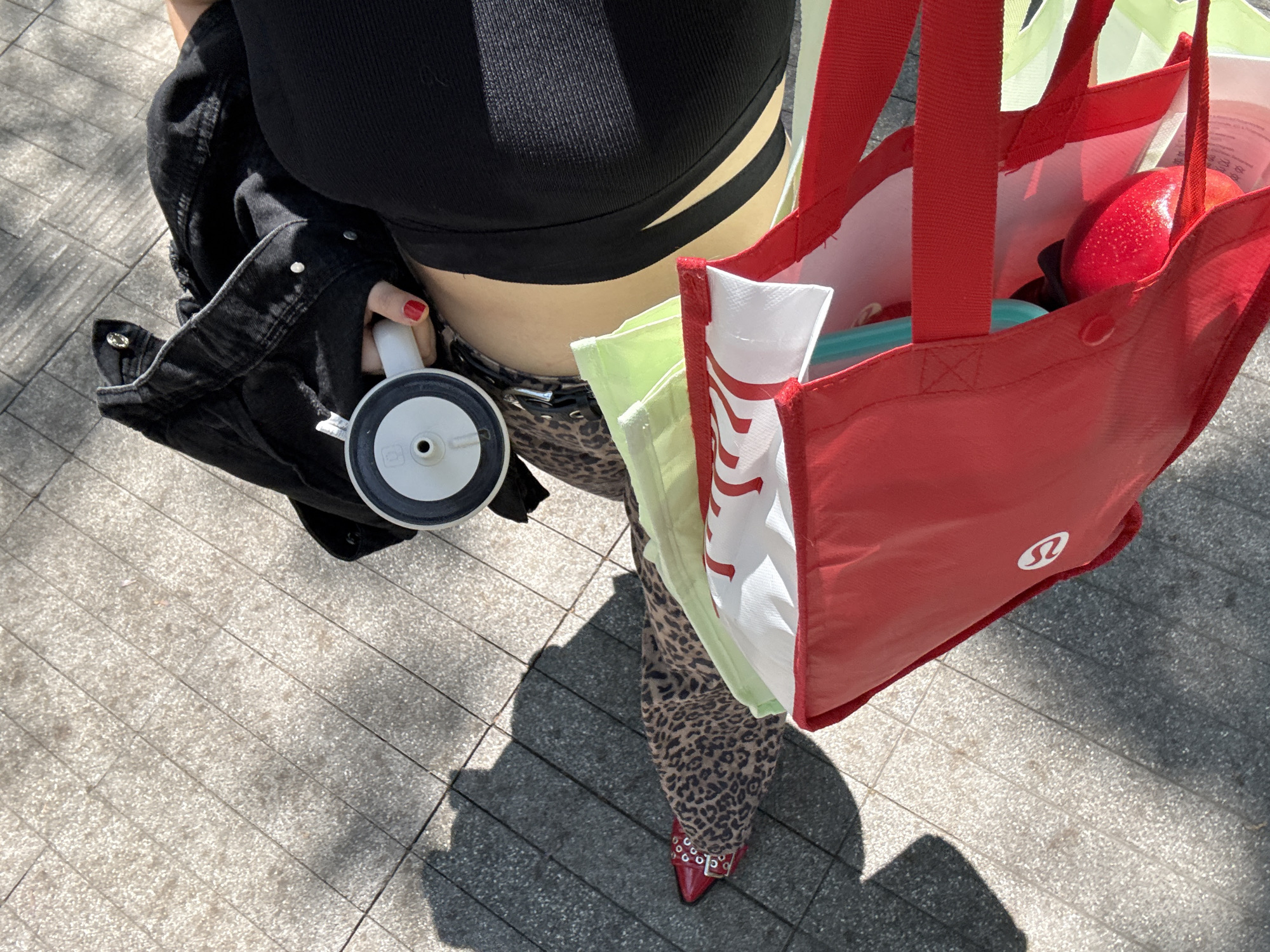 The image captures a high-angle, close-up view of a person from the waist down, walking outdoors on a sunny day. The person is wearing a black ribbed crop top with cut-out details on the midriff, a black denim jacket slung over one arm, and leopard-print pants. On their feet are distinctive red pointed shoes with silver stud embellishments and a buckle strap.
The person holds a white cup with a black and white lid in one hand, featuring a central hole. The other hand carries several reusable shopping bags. The prominent bag is red, made of a fabric-like material, and features the white "LULULEMON" logo on its side. Inside this red bag, a glossy red plastic apple or fruit is visible, along with glimpses of other items. Behind the red bag, a light green reusable bag and a white one with red text (partially obscured) are also held.
The setting appears to be a paved urban area, possibly a sidewalk or pedestrian zone, with light-colored rectangular tiles. Strong sunlight casts distinct, dark shadows of the person and their belongings onto the ground, indicating a bright, clear day. The overall impression is of someone casually moving through an urban environment, possibly after shopping.