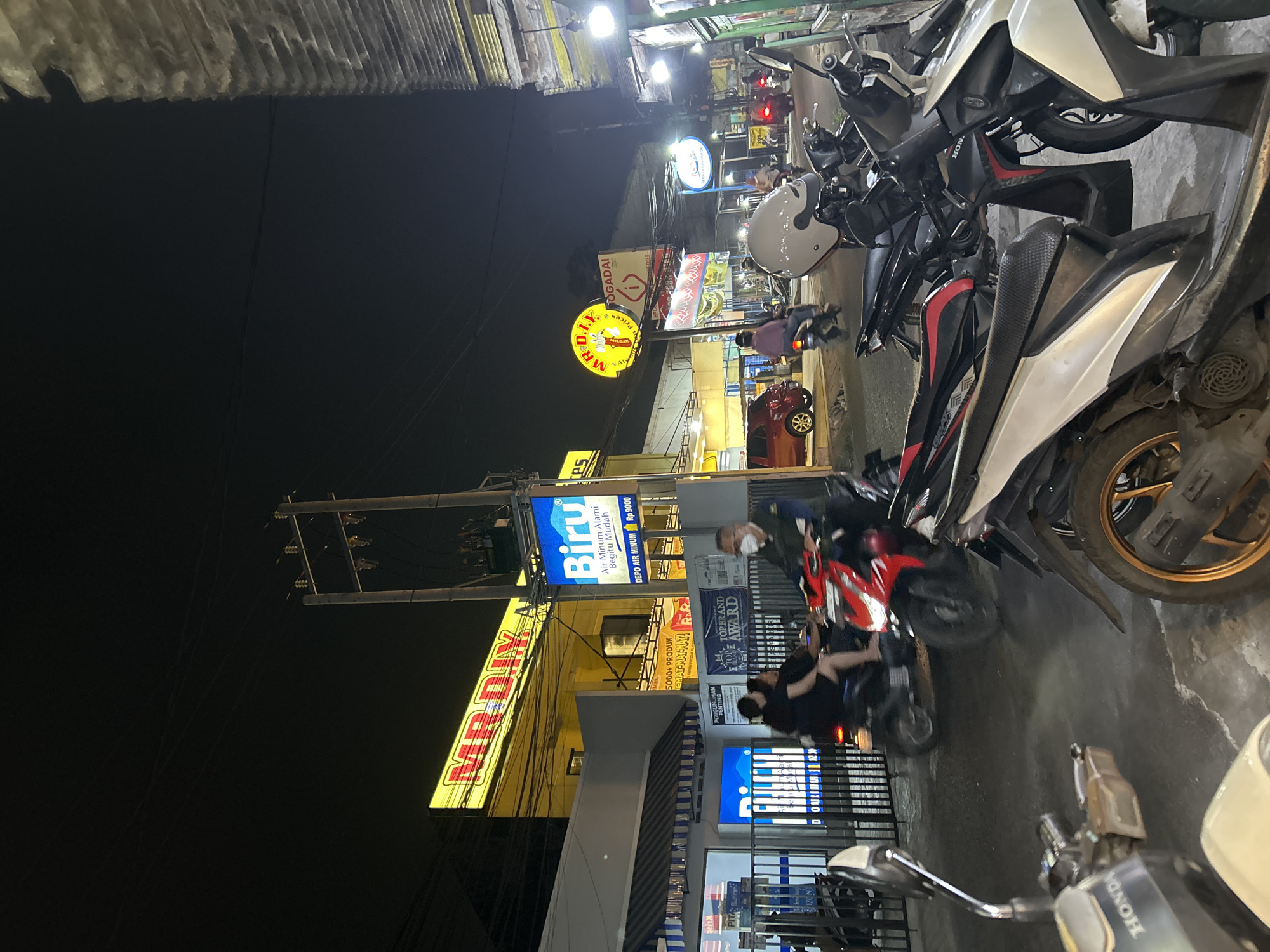 The image depicts a nighttime street scene in Depok, Indonesia, likely a commercial area with several businesses. Prominently displayed is a large, illuminated yellow sign for "MR. D.I.Y.", a retail chain. Adjacent to it is a blue sign advertising "Biru Air Minum Alami Begitu Mudah" (Blue Natural Drinking Water). A red car is parked on the right side of the street.
Several people are visible, primarily on motorcycles. Two individuals are riding a red motorcycle in the foreground, moving from left to right. Another person is seen riding a darker motorcycle further down the street. A man wearing a mask is walking or riding a motorcycle on the right side.
The street is lined with storefronts and illuminated signs. Motorcycles are parked along the sidewalk on the right side of the image, suggesting a common mode of transportation in the area. Power lines are visible crisscrossing above the street, a typical urban infrastructure element. The overall atmosphere is that of a busy commercial street at night, with artificial lighting from the businesses and vehicles illuminating the scene. The architecture appears to be a mix of modern and functional commercial buildings.