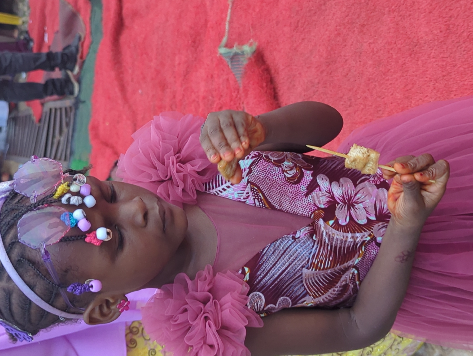 A young girl is depicted in a medium, eye-level shot, turned slightly to her left, with her head tilted towards the left, set against a vibrant reddish-pink background. Her skin tone is dark brown, and her hair is intricately braided, adorned with colorful beads and butterfly-shaped hair clips, some of which are translucent and iridescent. She is wearing a light pink headband. Her earlobes have small flower-shaped earrings, one purple and one pink.
She is dressed in a light pink dress with a ruffled, net-like material around the shoulders. The bodice features a burgundy fabric with a white floral and abstract pattern. Her hands are visible; the left hand is holding something that appears to be food, possibly a piece of fried yam or tofu, on a wooden skewer. Both hands show hints of an orange or red tint on the fingertips, possibly from henna or food coloring. She has a faint tattoo or mark on her right forearm.
In the background, on the far left, a person's legs and feet are visible, clad in dark pants and shoes. A dark wooden or metal railing is also partially visible behind them. The reddish-pink background appears to be a textured fabric, possibly a carpet or blanket, draped over a surface. Above the girl's right hand, a light-colored, possibly white, string or rope with a green and white tassel-like object is hanging down from the top edge of the frame. The lighting suggests it is daytime. The image captures a moment of focus or contemplation as the girl looks down at what she is holding.