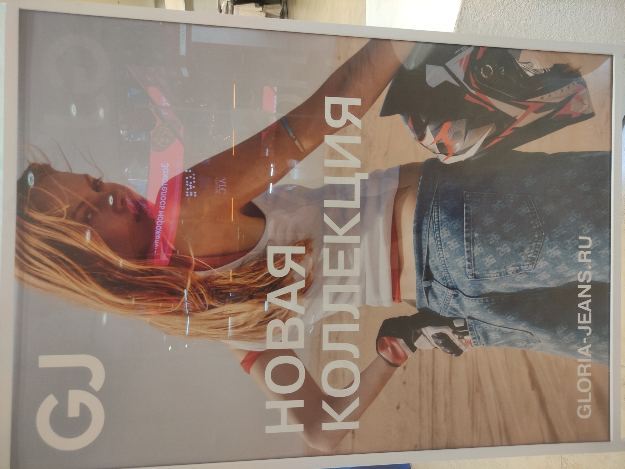 The image shows a promotional poster for Gloria Jeans, a clothing brand, in what appears to be a shopping mall or store in Moscow, Russia. The poster depicts a woman with blonde hair wearing a white top with red trim, a denim bottom (likely jeans), a motorcycle helmet, and gloves. The text "НОВАЯ КОЛЛЕКЦИЯ" (New Collection) is vertically oriented on the poster along with the brand name, "GLORIA-JEANS.RU", also vertically oriented. The initials "GJ" in a stylized font are visible on the left side of the poster. Reflections in the glass cover of the poster suggest the surrounding environment is indoors, probably within a shopping center due to the visible store details and lighting. The image captures a typical retail advertisement scene.