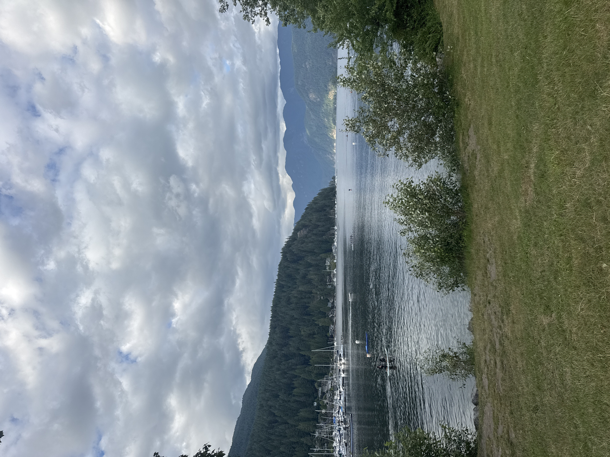 The image depicts a scenic view of Deep Cove in the District of North Vancouver, Canada. The foreground features a grassy area with some bushes. The middle ground shows a calm body of water, likely a bay or inlet, with several people engaged in water activities. Two individuals are visible on a paddleboard, and a small boat is moving across the water. In the background, a marina with numerous sailboats is docked along the shoreline, with a densely forested mountain slope rising behind it. The mountains continue in the distance, fading into a hazy blue against a cloudy sky. The overall atmosphere is serene, with the water reflecting the overcast sky and the lush greenery of the surrounding landscape.