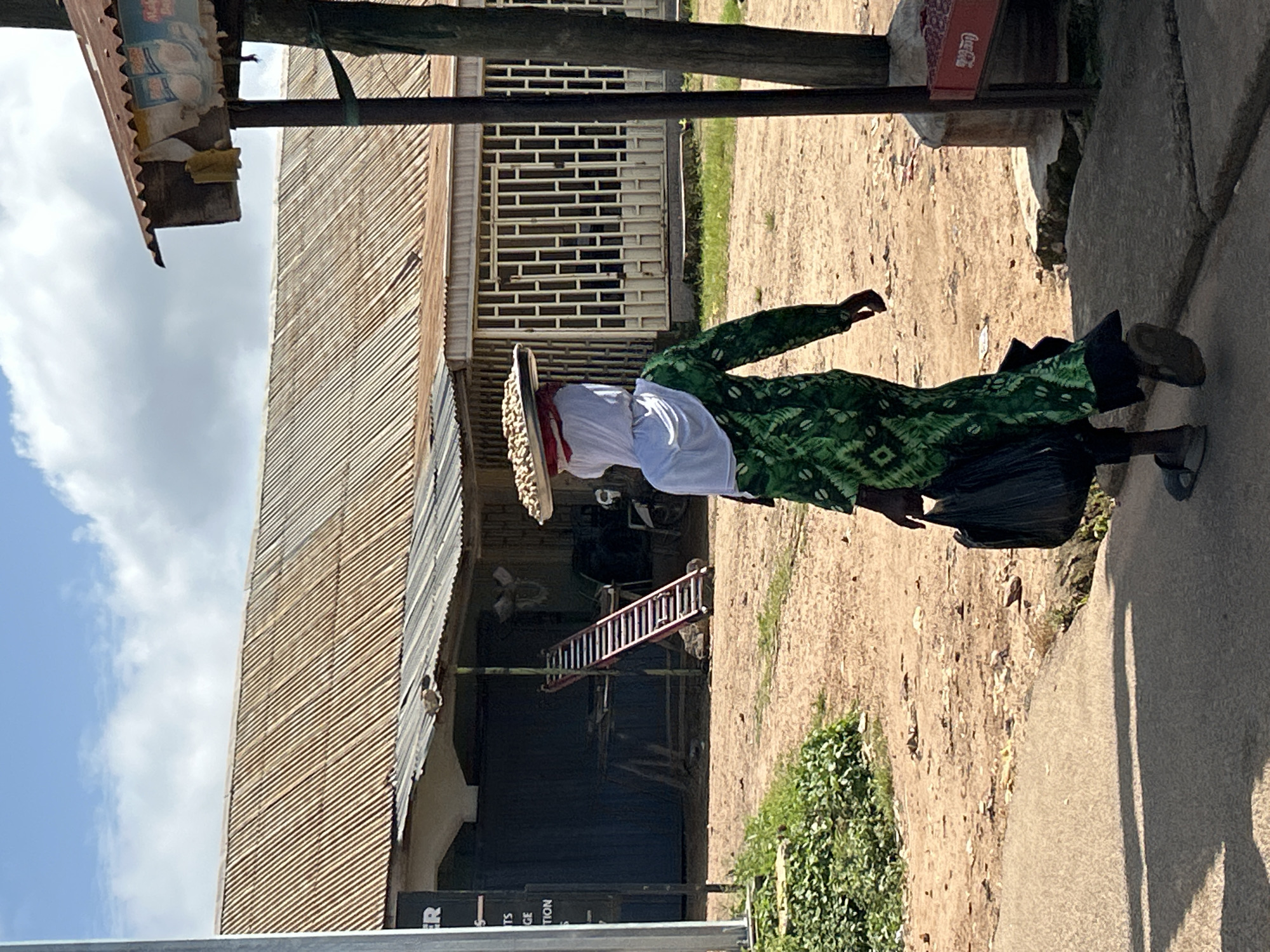 In Jos, Nigeria, a woman walks away from the camera on a dirt path. She wears a green, patterned dress and a white headscarf. On her head, she balances a round tray filled with light-colored nuts or seeds. In her left hand, she carries a black plastic bag. The background features a corrugated metal roof extending over a building with barred windows. A red ladder leans against the side of another structure. A wooden post with a Coca-Cola advertisement attached stands to the right of the frame. The sky above is partly cloudy.