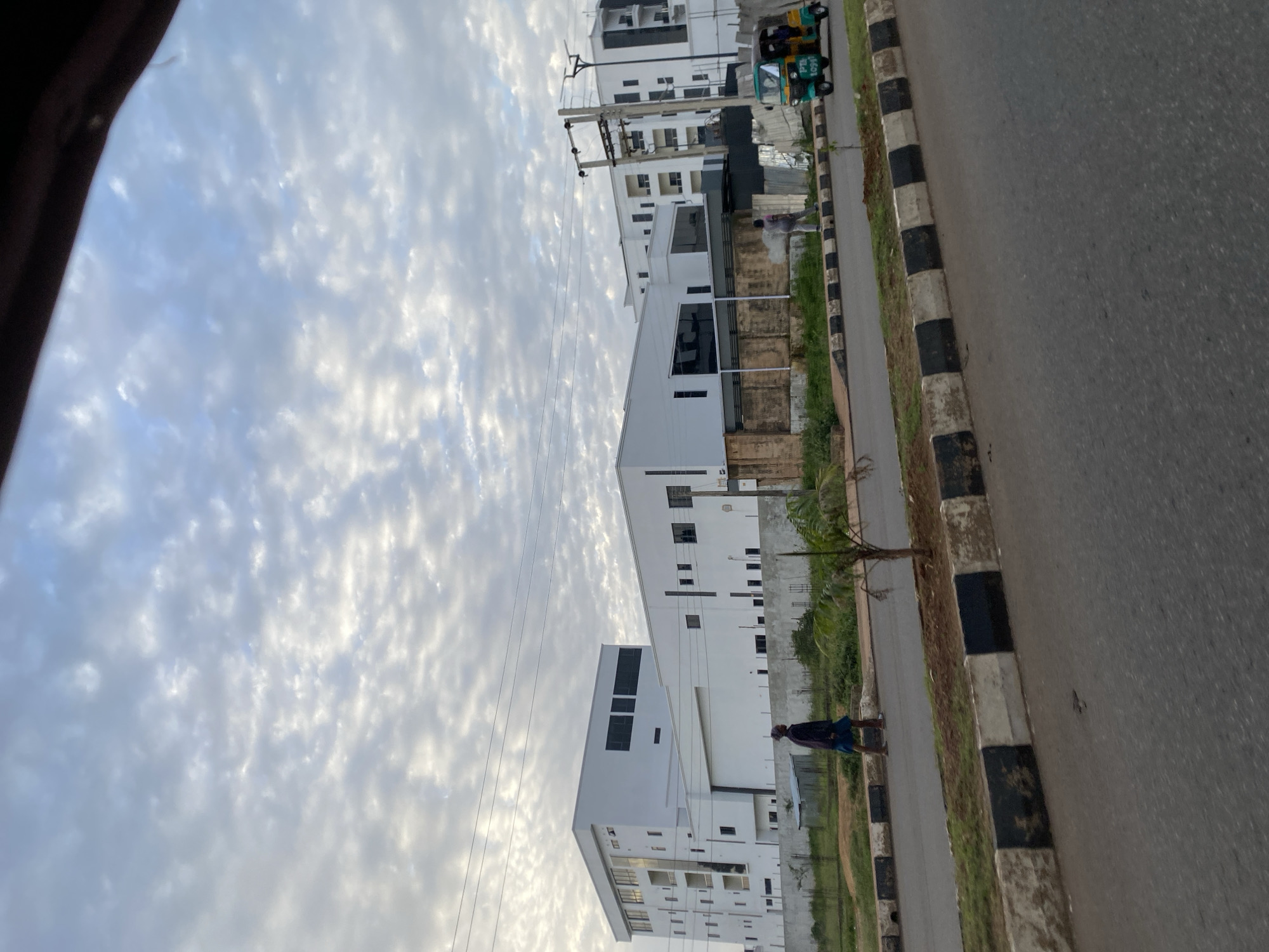 The image shows a street scene in Bar Jirgi Summa, Nigeria, with modern white buildings in the background. A person in dark clothing is walking along the side of the road, and a three-wheeled vehicle, commonly known as a Keke Napep, is also visible. The sky is partly cloudy, with a mix of white and gray clouds. The road is paved and has a distinct black and white striped curb. The area appears to be an urban or semi-urban environment.