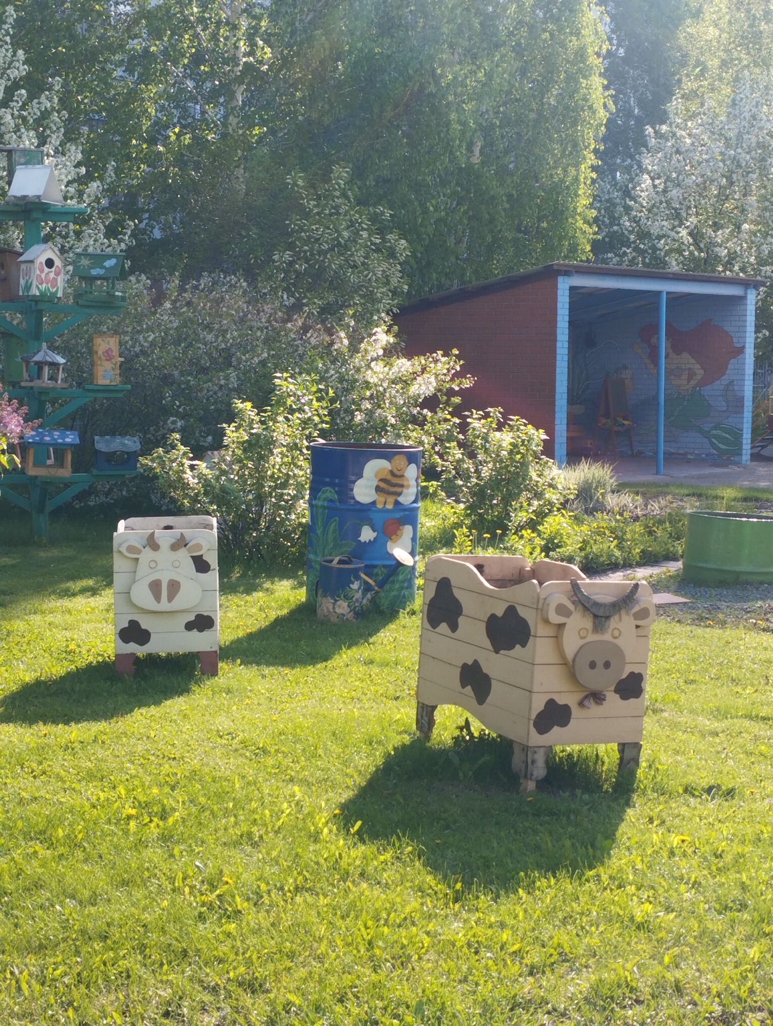 The image shows an outdoor scene, likely a park or garden, with several whimsical, hand-painted decorations. Two cow-shaped planters sit on a grassy lawn, one light in color with dark spots and the other white with dark spots. In the background, a painted barrel with a bee and flower motif can be seen.
To the left, a multi-tiered birdhouse structure stands tall. Behind the barrel, there is a small brick building with a blue trim, and a mural visible inside the covered area. The area is overgrown with bushes and flowering plants. The location is in the city of Novosibirsk, Russia.