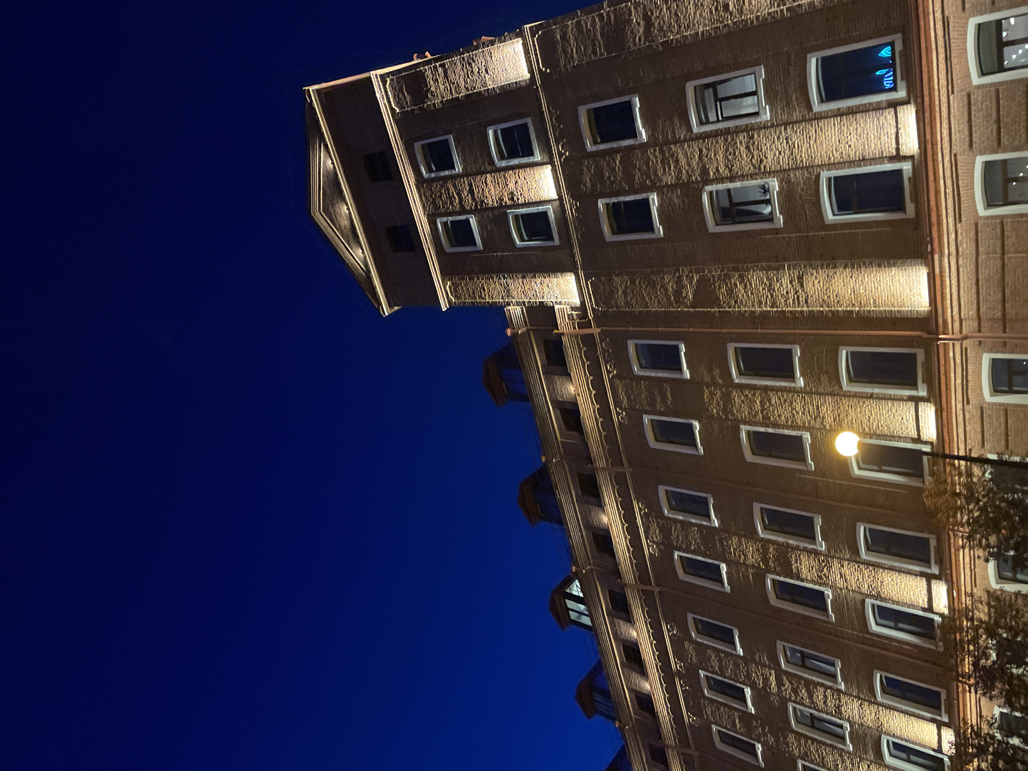 A low-angle shot depicts a multi-story brick building against a deep blue night sky. The building appears to be located at 15 Malysheva Street in Yekaterinburg, Russia. The brick facade is illuminated by warm lighting, highlighting rows of rectangular windows. A lamppost stands near the building, adding to the illumination. The architecture of the building is classic with ornate design elements along the edges. The building is angled, with the left side higher than the right, creating a dynamic perspective. The contrast between the warm light on the brick and the deep blue sky is significant.