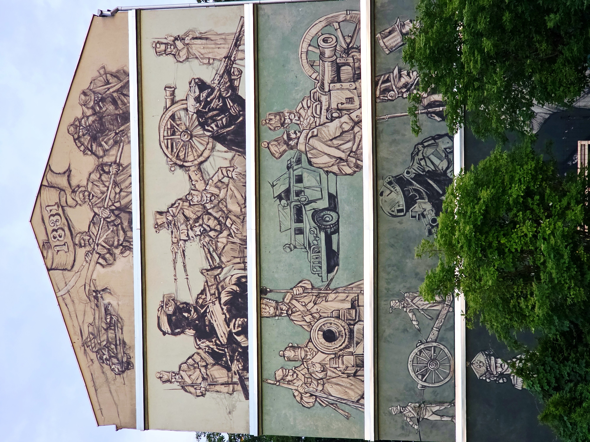 This is a mural painted on the side of a building in Warsaw, Poland, depicting historical and modern military scenes. The mural is divided into four horizontal sections.
The top section, painted on the gable end of the building, features a banner with the number "1831" and shows a group of soldiers in historical uniforms preparing to pull a cart. The style is a rough sketch.
The second section from the top shows a line of soldiers in historical uniforms, some holding rifles and bayonets, and others operating a large cannon. In the background, a modern military vehicle resembling an armored personnel carrier is visible.
The third section from the top depicts a group of soldiers in historical uniforms gathered around a cannon. Another modern military vehicle is parked to their right.
The bottom section shows a modern soldier wearing a helmet and tactical gear, holding a rifle, in the foreground. Behind this soldier are figures in historical military uniforms, one of whom is holding a rifle. The lower part of this section is partially obscured by lush green trees. The overall style of the mural is a sketch with bold lines and shading, predominantly in black, white, and grey tones against a muted background.