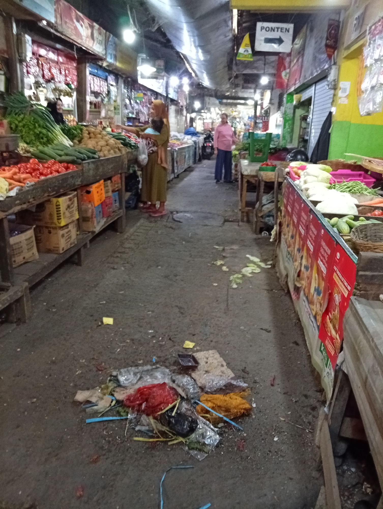 The image depicts an indoor market, likely in Larangan, Indonesia, with a focus on the pathway and stalls. The ground is concrete and appears dirty, with scattered debris including plastic bags, food scraps, and some yellow paper. To the left, a stall displays an abundance of fresh produce, including tomatoes, carrots, cucumbers, and green onions. Boxes are stacked beneath the stall. Further down the pathway, a woman in a headscarf is seen near the produce stall, appearing to be interacting with customers or managing the merchandise. In the background, another woman in a pink shirt and blue pants stands near a green plastic crate. A sign with the word "PONTEN" and an arrow pointing right is visible overhead. To the right, another stall is laden with various vegetables, including what appear to be cabbage, cucumbers, and green beans, with promotional banners displayed on its front. The overall scene suggests a busy marketplace environment with an ongoing transaction of goods.