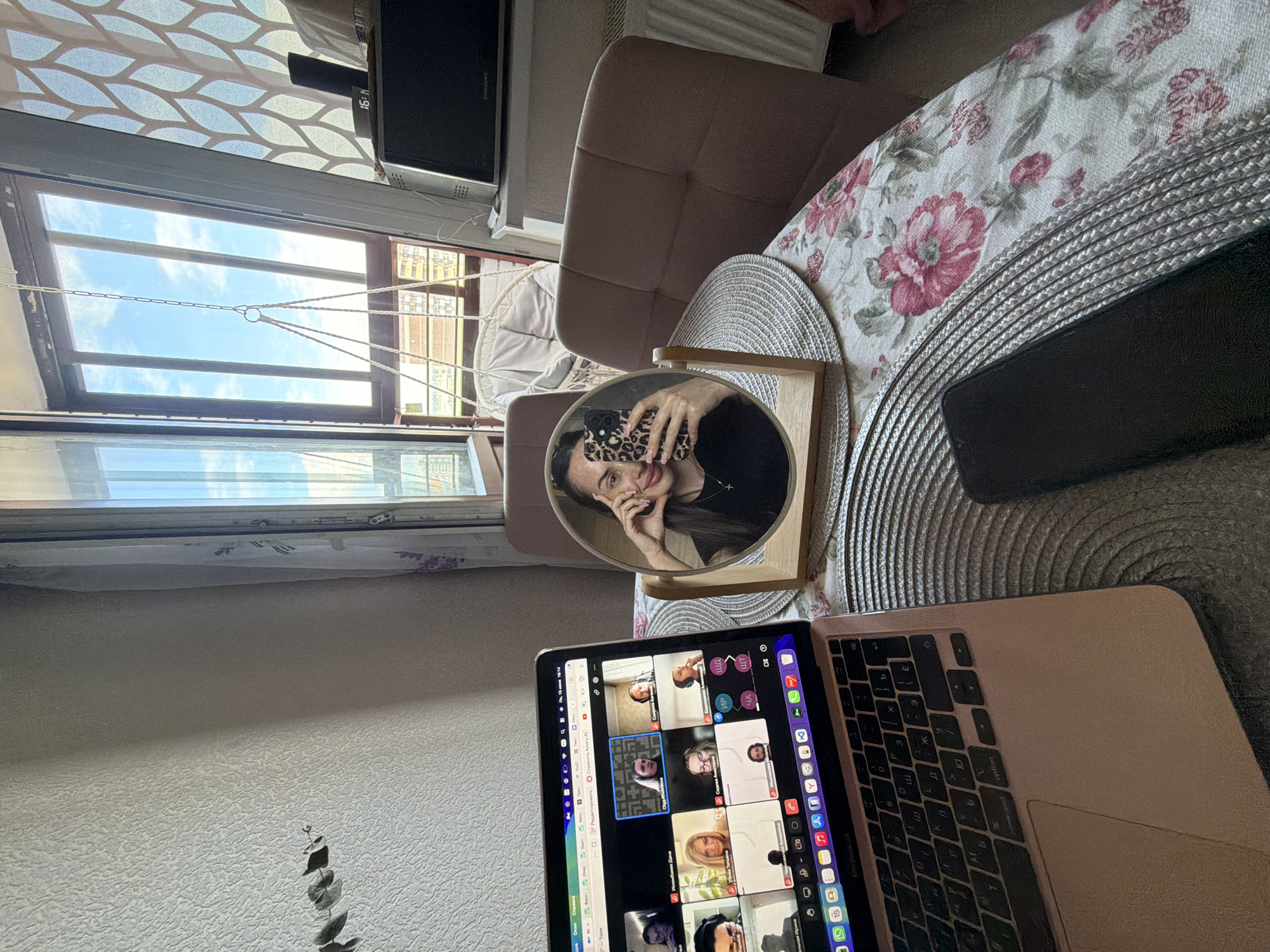 The image depicts a young woman taking a selfie in a round mirror placed on a table covered with a floral tablecloth. She is seated indoors, possibly in an apartment, with a laptop open in front of her, displaying a video conference with multiple participants. The background reveals a window with a view of a balcony and the sky, suggesting an urban setting. A hanging chair is visible on the balcony. The overall scene conveys a sense of a work-from-home or remote learning environment. No specific details to indicate the location in Saint Petersburg, Russia.