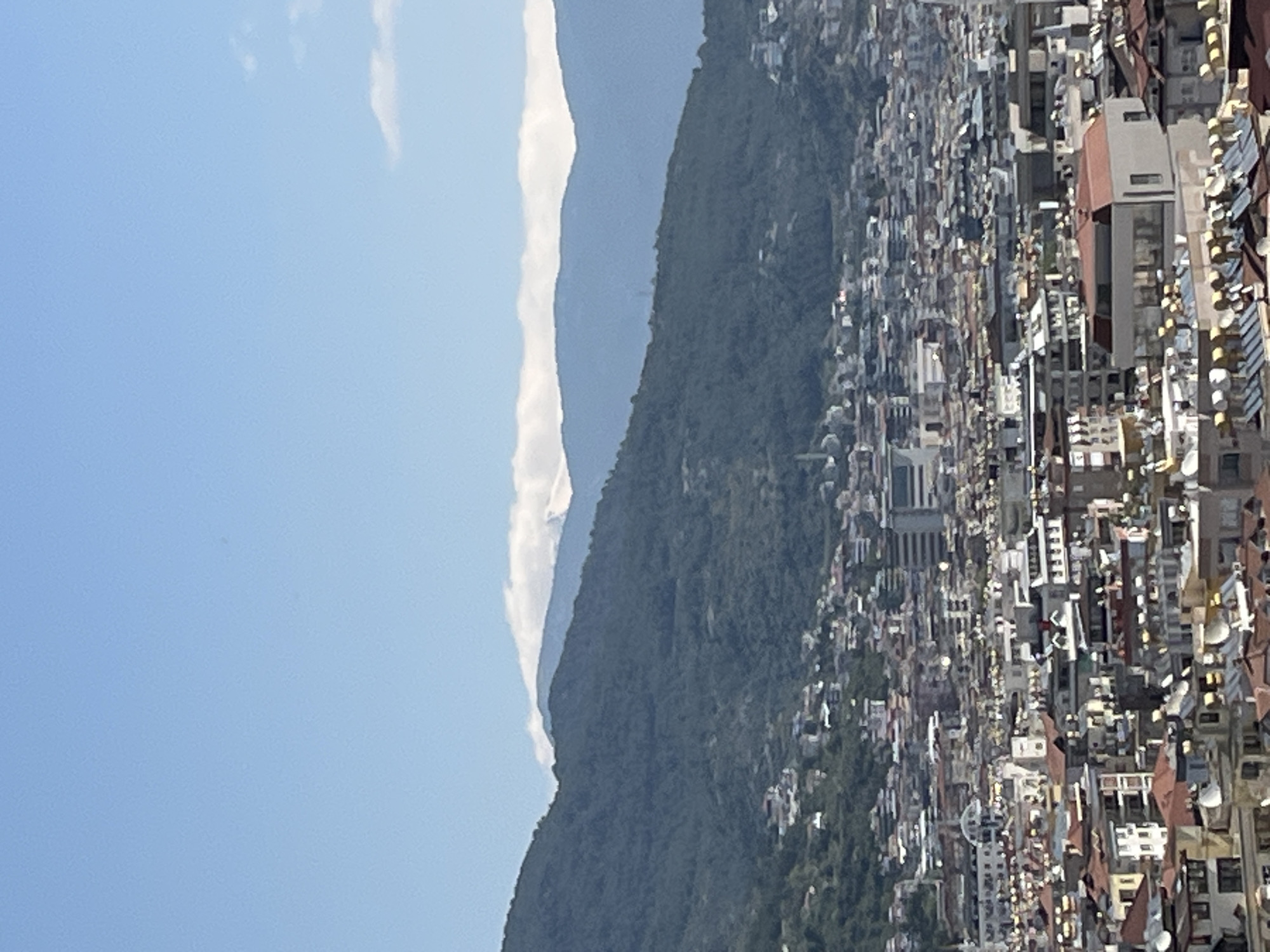 The image shows a view of Alanya, Turkey, from a high vantage point. In the foreground is a dense urban landscape with buildings of varying heights and architectural styles. The rooftops are predominantly white and terracotta. Behind the city, a verdant mountain range rises, its slopes covered in trees and vegetation. A strip of white clouds is visible next to the mountain. The sky above is clear and blue.