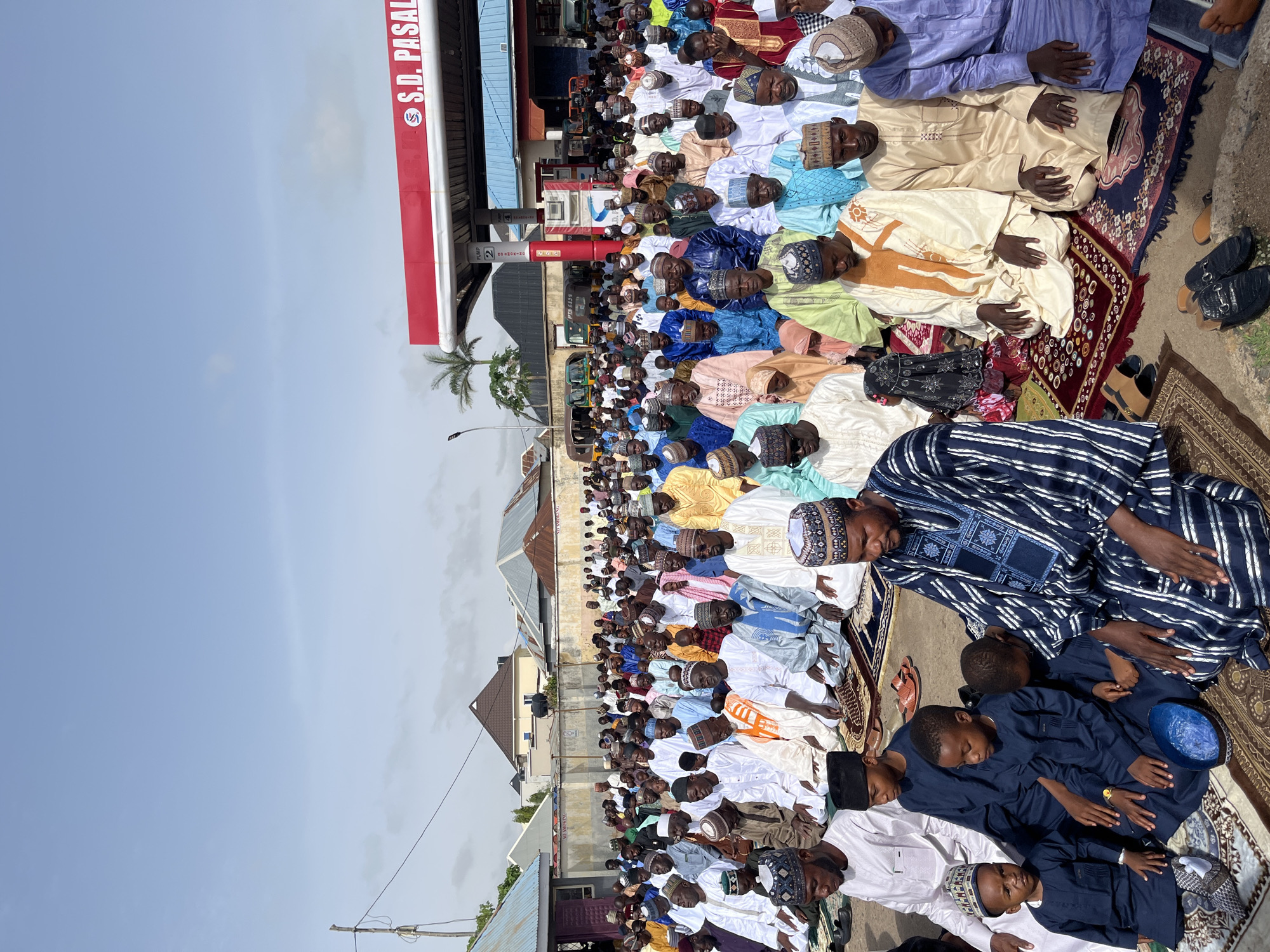 A high-angle, medium shot shows a large group of people kneeling and praying in front of a gas station. The gas station is branded "S.D. PASAI." A group of men and boys, clad in colorful clothing and traditional caps, are arranged in rows on prayer mats. The location appears to be an urban area with residential buildings in the background. The sky is a pale blue. The city and country are unknown.