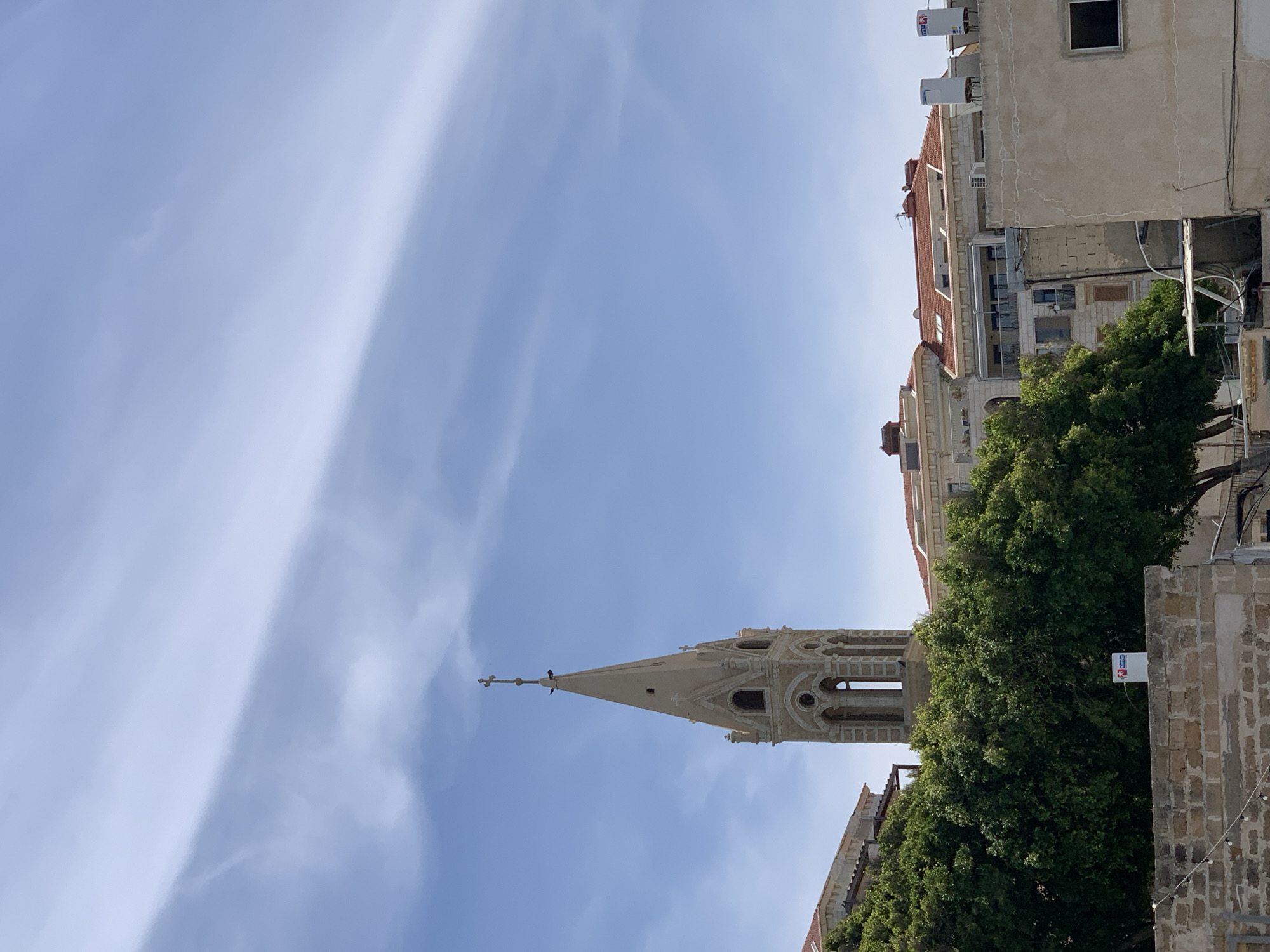 The image captures an upward view of an urban landscape in Tel-Aviv, Israel, under a bright, clear blue sky adorned with wispy white clouds. Dominating the central-right portion of the frame is a tall, light-colored church steeple. The steeple features ornate architectural details, including arched openings and decorative carvings, and is topped with a cross on which a small dark bird is perched.
To the right, a large, dense green tree obscures part of the steeple's base and several multi-story buildings. These buildings, stretching into the background on the right side of the image, display a mix of architectural styles. Some feature traditional red-tiled roofs, while others have lighter, flat facades, with visible modern elements such as water heaters on their rooftops. One building's wall shows cracked plaster, indicating age.
The scene depicts a peaceful daytime setting with abundant natural light, suggesting it is late morning or early afternoon. The weather appears clear and sunny. No human activity is visible, contributing to a sense of urban tranquility. A small white rectangular sign with red and blue accents is partially visible below the tree on the right, but any specific text on it is unreadable due to its size and distance. The overall image showcases a blend of historical architecture and modern urban life.
