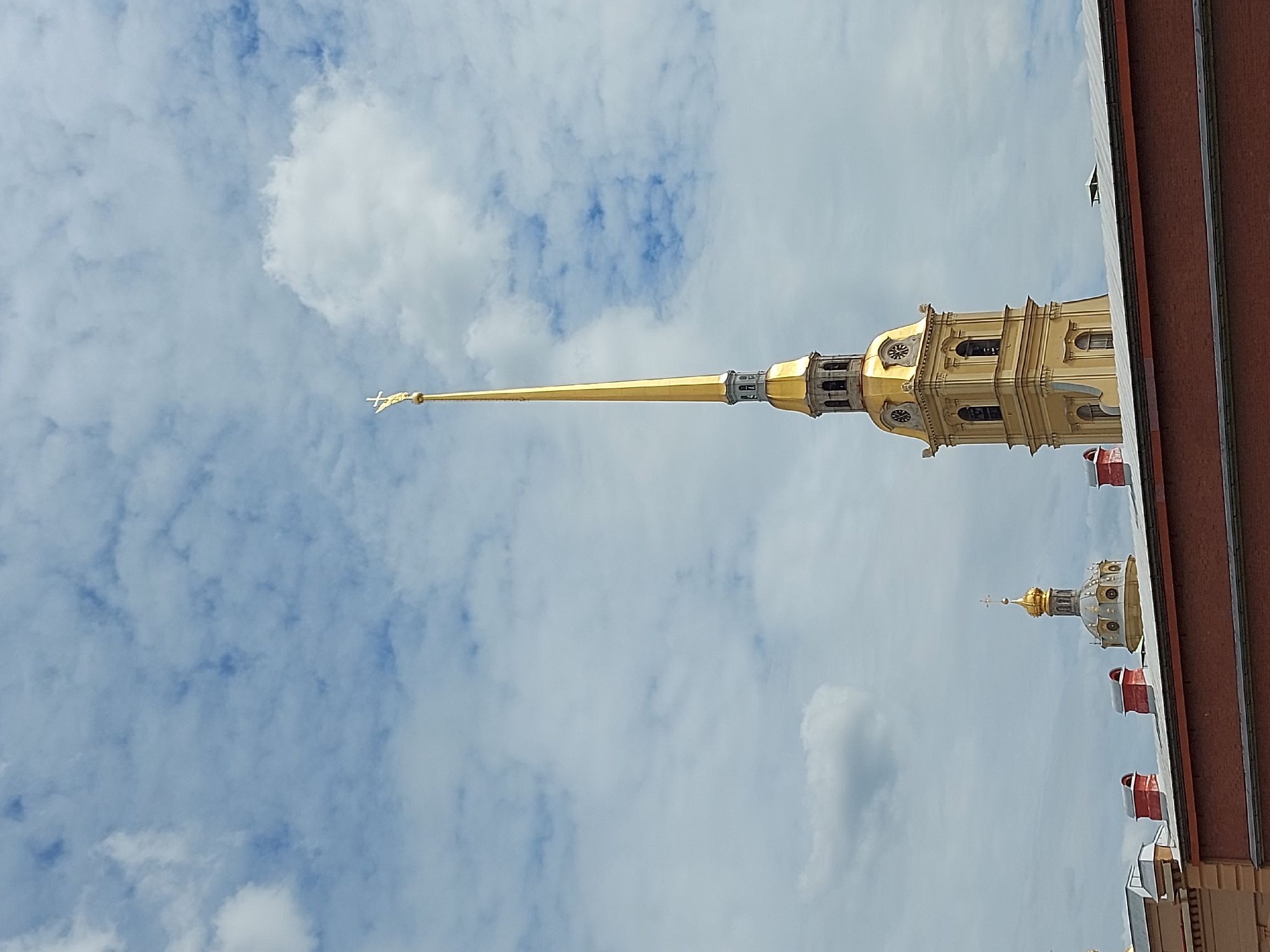 The image shows a spire with a golden needle against a cloudy sky. The spire is the Peter and Paul Cathedral in Saint Petersburg, Russia. The cathedral tower is visible on the right side of the spire, with clocks and windows visible on its golden facade. A second, smaller golden tower is visible in the bottom right corner, with red sections. The edge of a building is visible on the far right. The sky is blue with white and grey clouds.