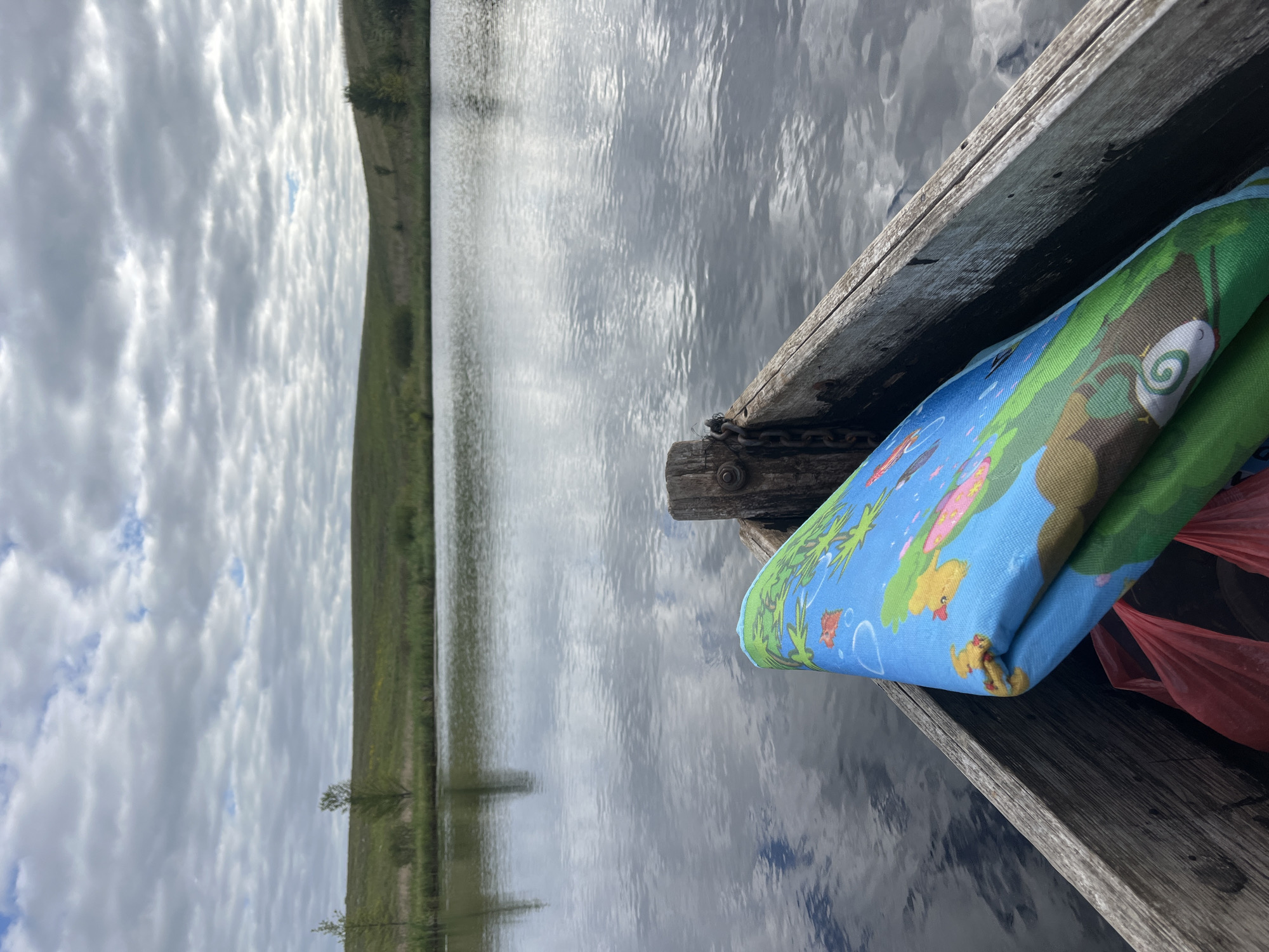 A first-person perspective shot from inside a weathered wooden boat shows a calm body of water with reflections of a cloudy sky. In the foreground of the boat, a rolled-up blue mat with a cartoonish world map design is visible, along with a red plastic bag. The background features rolling green hills sparsely dotted with trees, indicative of a rural landscape. The overall atmosphere is tranquil and natural. The location is in the vicinity of Дунілавіцкі сельскі Савет, Belarus.