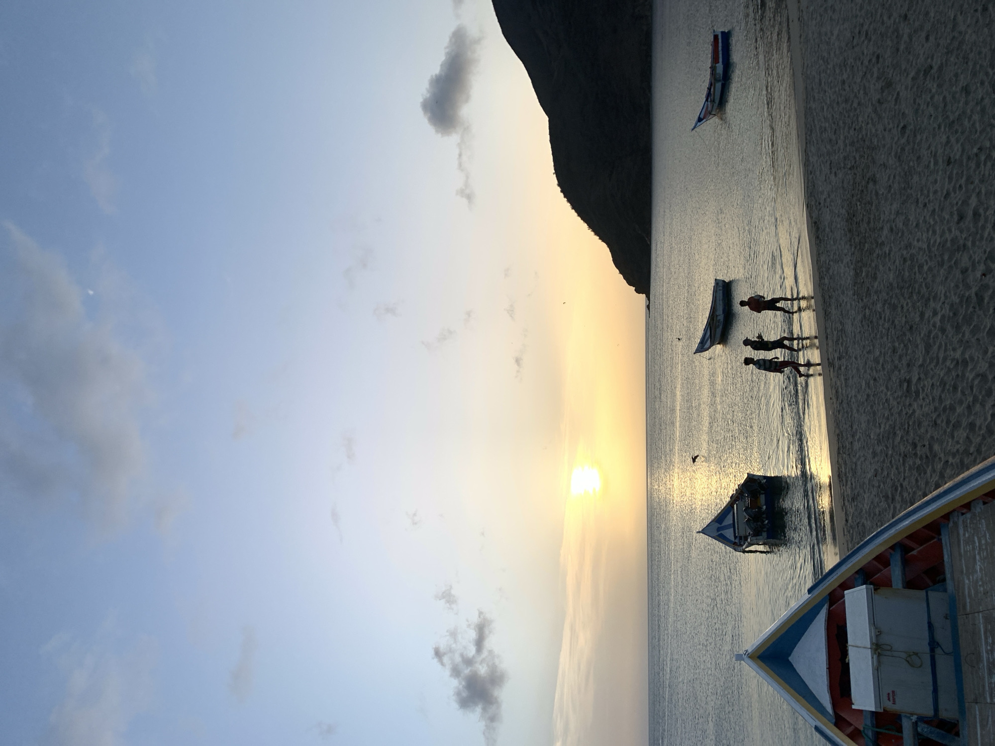 The image depicts a serene sunset scene on a beach in Juan Griego, Venezuela. The sky is a gradient of pale blue and soft yellow, with scattered clouds. The sun, a bright orb, is low on the horizon, casting a warm glow across the tranquil water. In the foreground, a colorful boat with a blue, yellow, and red trim rests on the sand. Several other boats are anchored in the shallow water. Three figures, silhouetted against the fading light, walk along the shoreline, their forms indistinct. In the distance, a dark, mountainous landmass rises from the sea, its slopes covered in vegetation. The overall atmosphere is one of peace and natural beauty.
