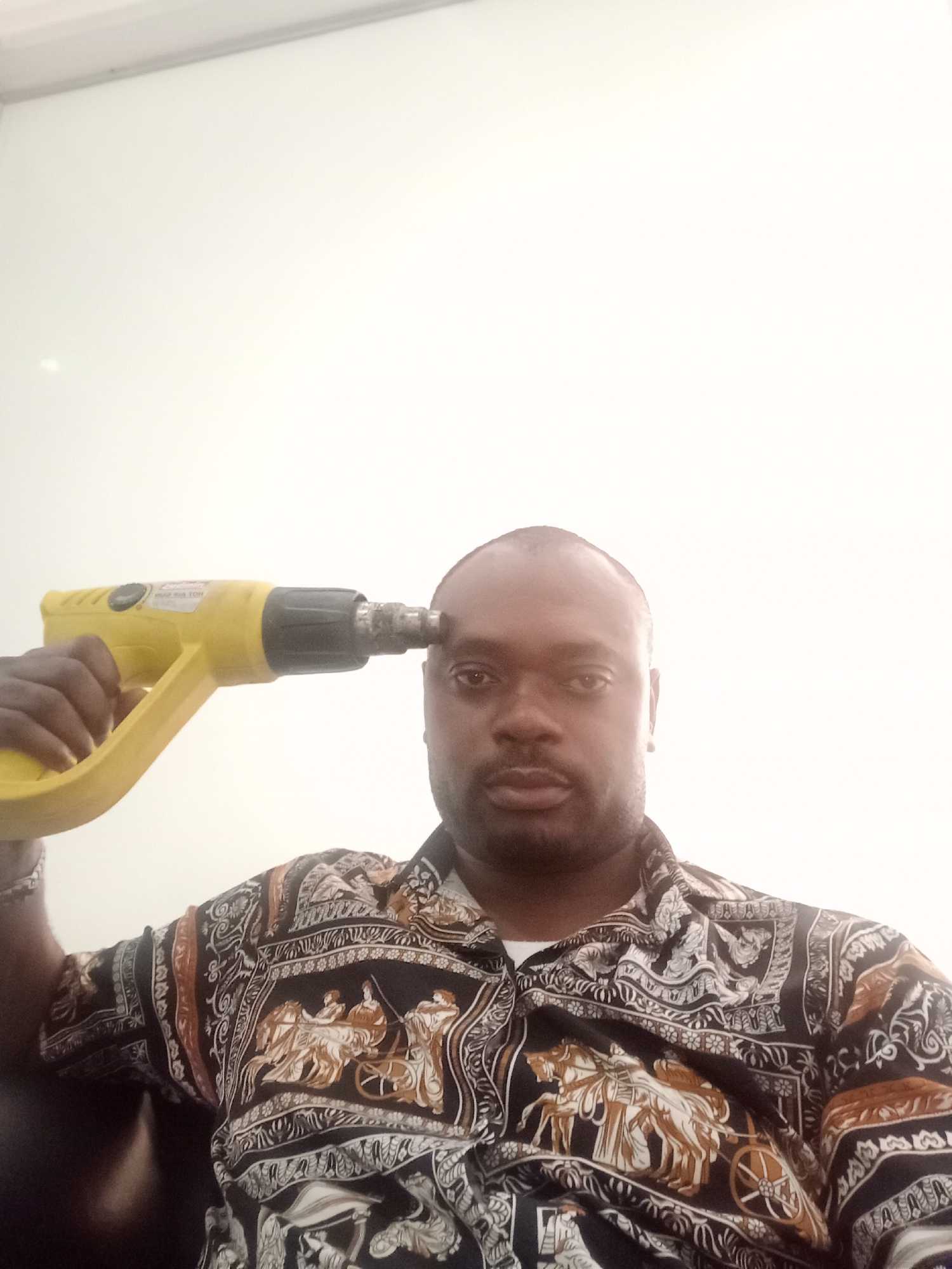 An indoor, medium shot captures a Black man holding a yellow heat gun to his right temple. He looks directly forward with a neutral to serious expression. The man has a closely shaven head and wears a dark, short-sleeved collared shirt. The shirt features an intricate pattern of classical or historical scenes, depicting figures riding in chariots drawn by horses, interspersed with decorative scrolls and borders in shades of brown, white, and black on a dark background.
The yellow heat gun has a black nozzle and handle grip, resembling a pistol in shape. Partially visible, reversed text on the side of the tool appears to read "HOT AIR TOOL." The background is a plain, brightly lit white wall, suggesting an interior setting within Gwarinpa, Nigeria. The overall lighting is very bright, especially in the background.