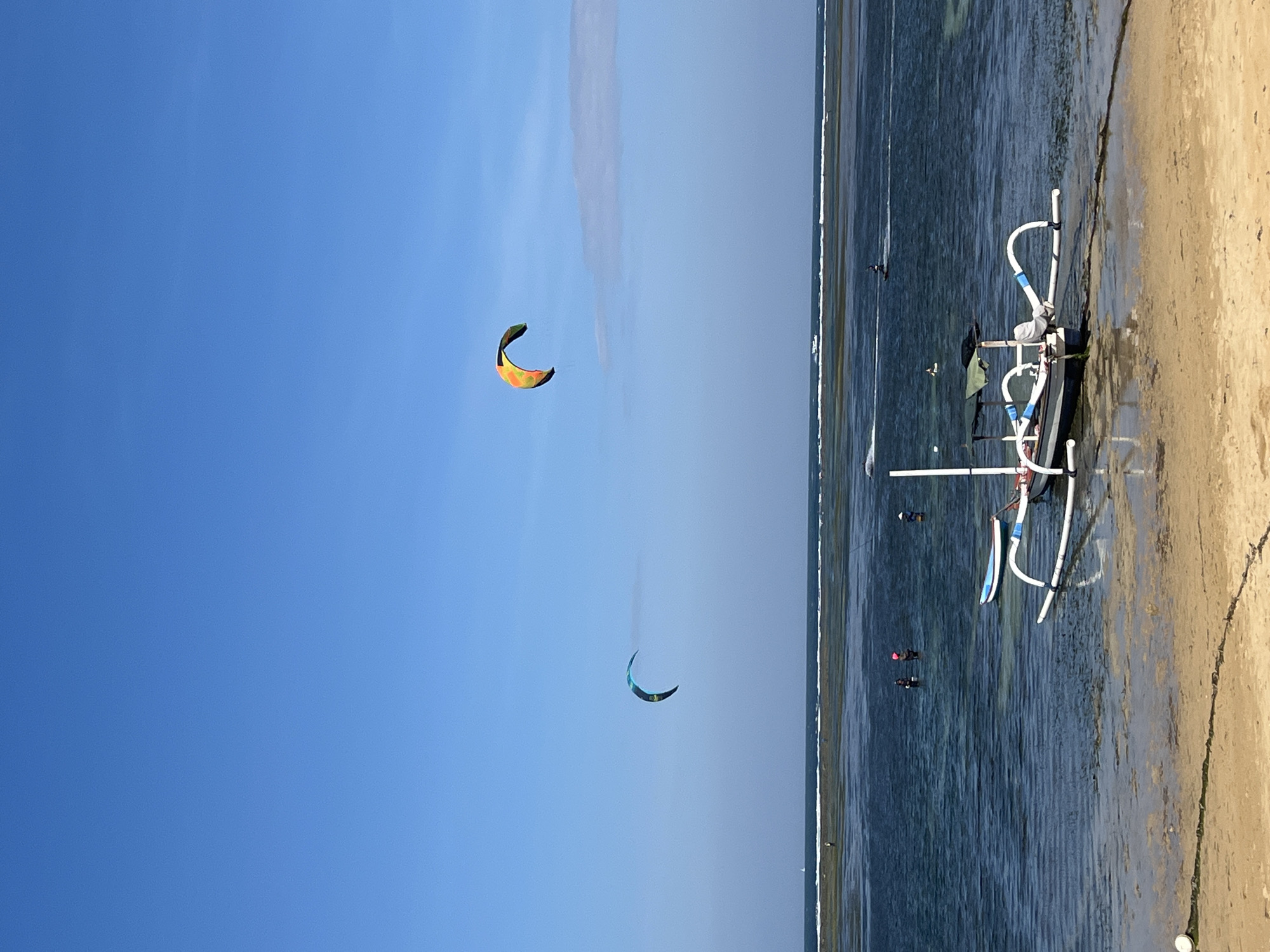 The image depicts a bright, sunny day at a beach in Denpasar, Indonesia, likely late morning or early afternoon given the clear blue sky and the angle of the sun creating distinct shadows. The scene is a vibrant mix of sky, sea, and sand. Two colorful kitesurfing kites, one orange and yellow, and another teal and black, are prominently displayed against the vast, clear blue sky, indicating kitesurfing is taking place.
In the shallow, clear blue water, several people are visible, some appearing to be kitesurfing or preparing to do so, as they are spread out across the water. A traditional outrigger boat, with white and blue accents, is partially submerged in the water near the shore, its distinctive frame adding to the local flavor of the scene. The beach itself is a light, sandy color, with the gentle tide lapping at the shore. The overall mood is one of leisure and outdoor recreation, with the bright sunshine and active pursuits suggesting a pleasant day. There is no visible text in the image.