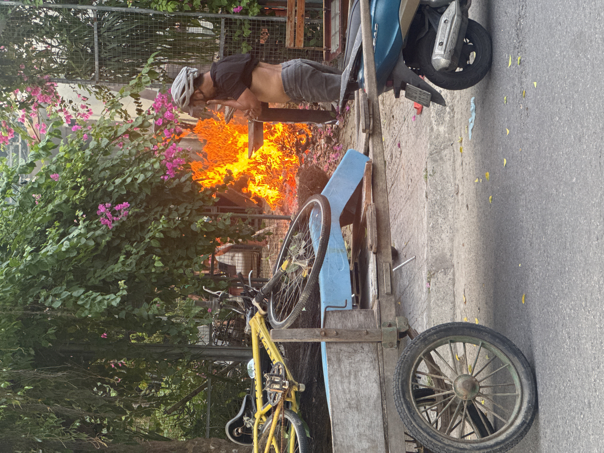 A horizontal photograph shows a man standing over a fire. He wears a bike helmet, a black shirt, and light-colored pants. The man has his head down, looking at the fire. The fire is burning in what appears to be a metal structure. To the right of the fire is a parked scooter. To the left of the fire is a wooden and blue bicycle trailer with a yellow bicycle attached. A large, green, flowering bush is behind the fire. The photo was taken outdoors during the day on a city street in Đà Nẵng, Vietnam.