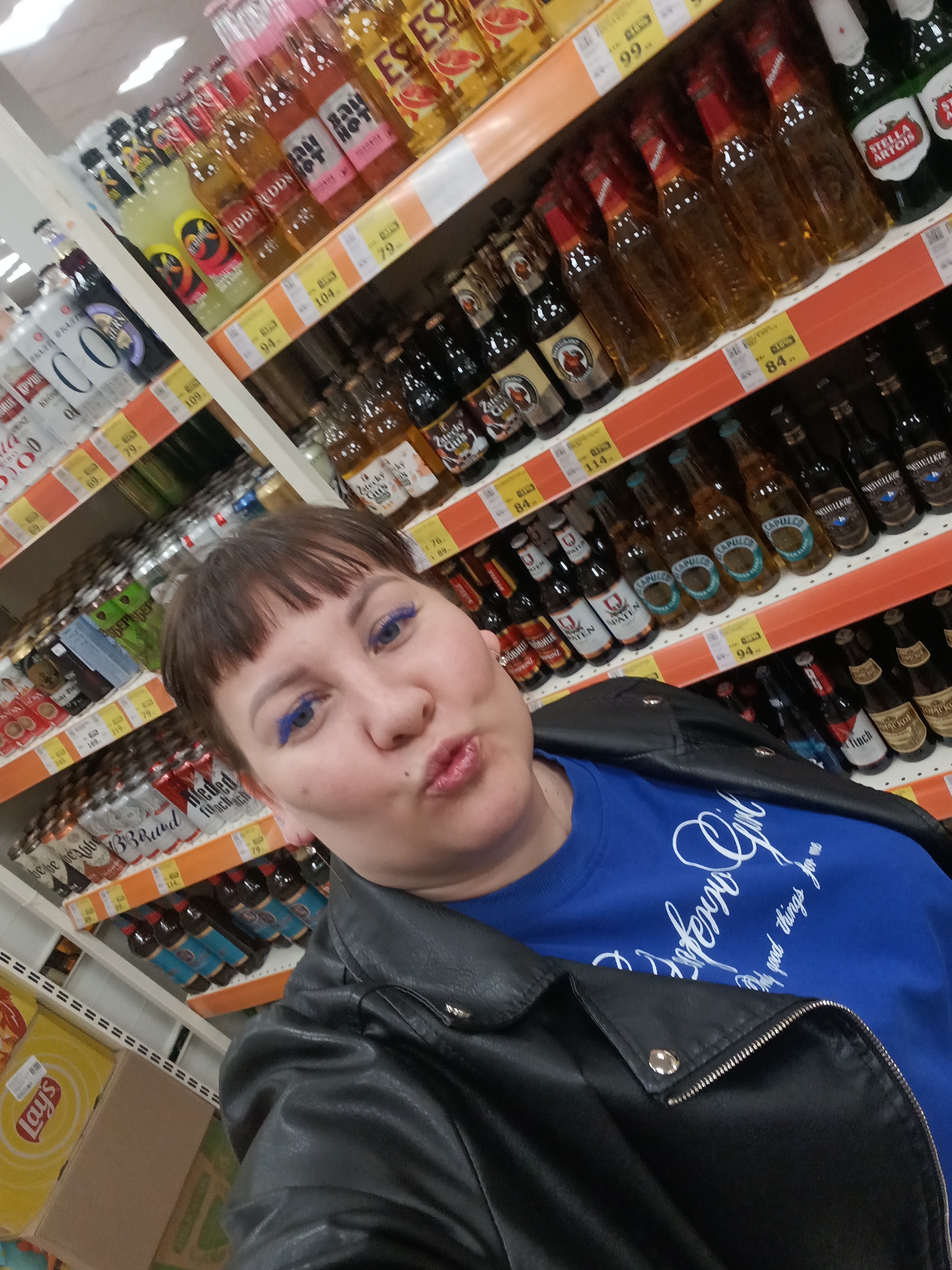 A woman with dark hair and bangs, wearing a blue sweater and a black leather jacket, takes a selfie in a grocery store. She has blue eyeliner and is making a kissing face. She stands in an aisle with shelves stocked with various alcoholic beverages. The shelves are labeled with prices in Russian rubles. Brands visible include Stella Artois, Zatecky Gus, and Eddy's. Boxes of Lay's chips are visible at the bottom of the frame. Based on the currency and brand availability, the location is in a Russian-speaking region, potentially Russia. There is not sufficient information to determine if the image was taken in the city of Osa, Russia.