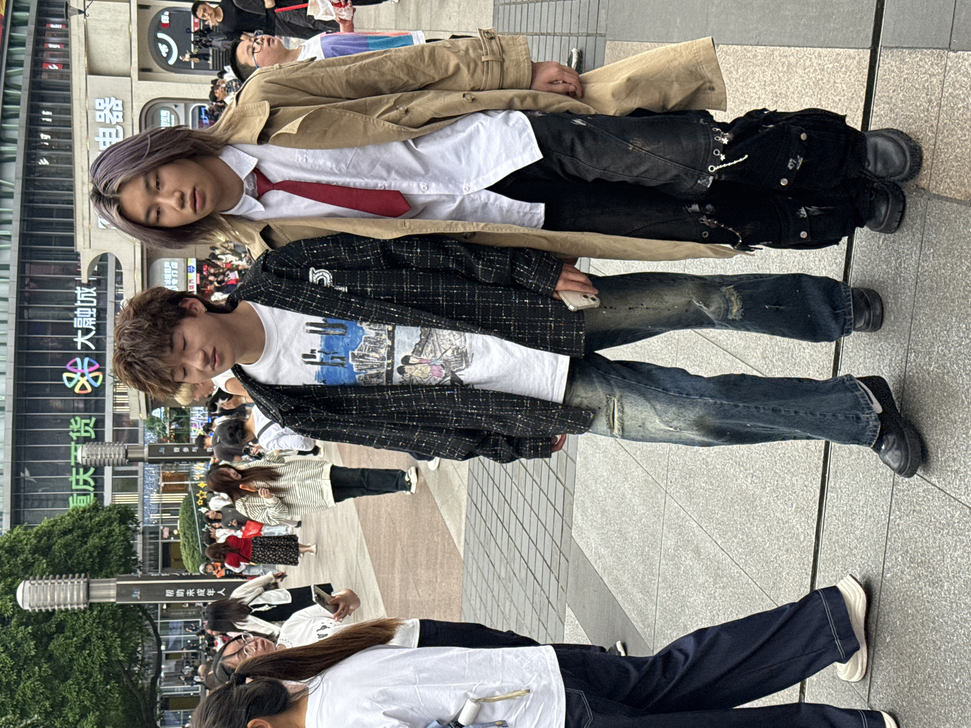 On a public pedestrian plaza in Liangjiang, China, two young individuals stand out amidst a larger group of people.
The first, an individual with short brown hair, wears a graphic t-shirt under a dark, checkered jacket, and distressed denim jeans. They are holding a smartphone.
Above, another person with light purple hair is seen. They wear a white shirt and red tie, partially covered by a tan trench coat, dark pants with metallic embellishments, and black shoes.
Behind them, a building with Chinese characters is visible. Other individuals can be seen walking in the background.