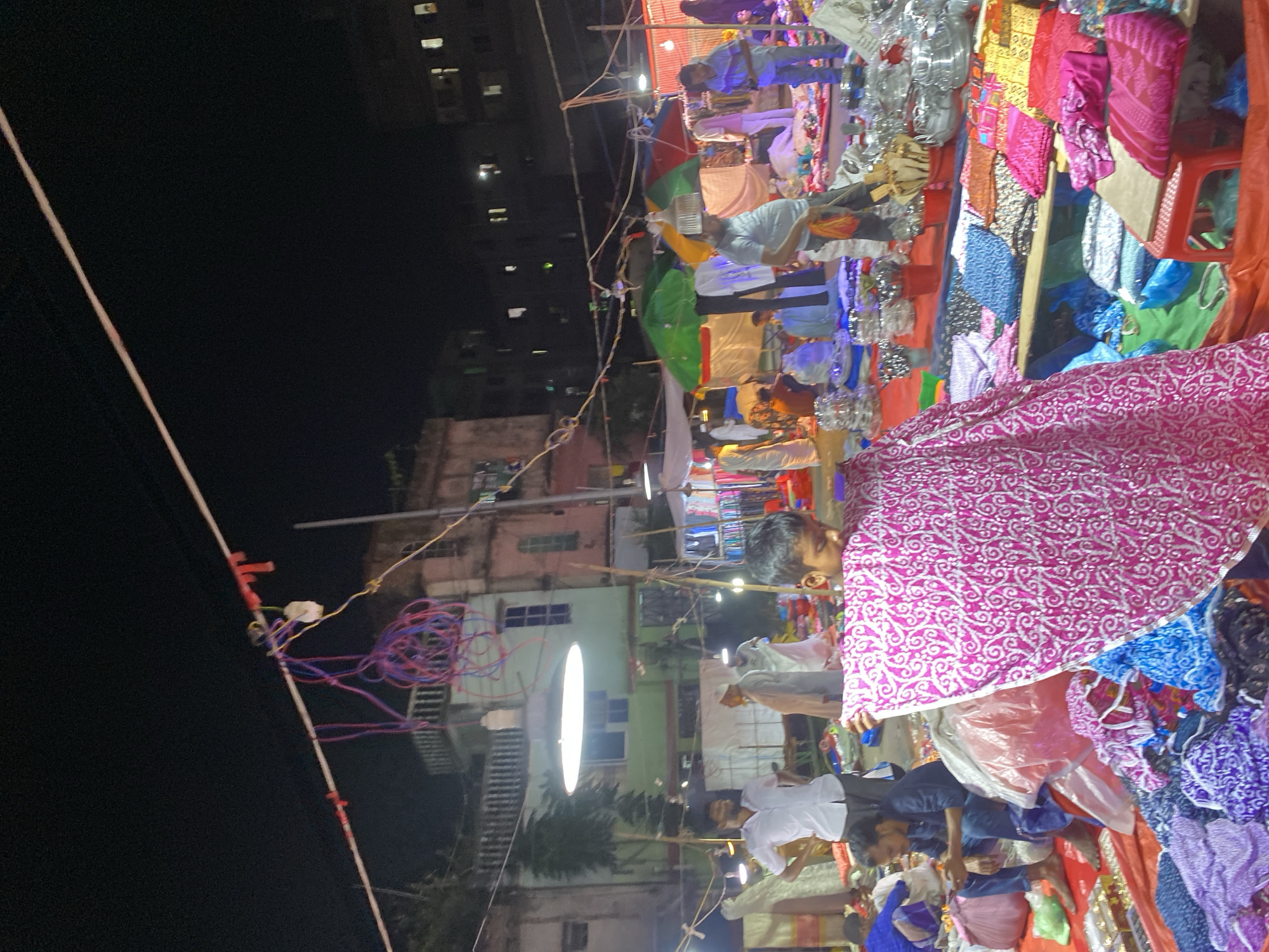 The image depicts a night market scene in Dhaka, Bangladesh, likely an outdoor bazaar. The foreground is dominated by piles of colorful textiles, including fabrics with intricate patterns in pink and white, and other vibrant hues. A young boy in the foreground is holding up a large piece of pink and white patterned fabric. In the mid-ground, several individuals are visible. A man in a white shirt stands near a stall, and further back, an older man in traditional attire is walking. The market appears to be set up under the open sky, with overhead wires and lighting fixtures illuminating the scene. Stalls with umbrellas are visible in the background, suggesting various vendors selling goods. The overall environment is bustling, indicative of a lively marketplace at night.