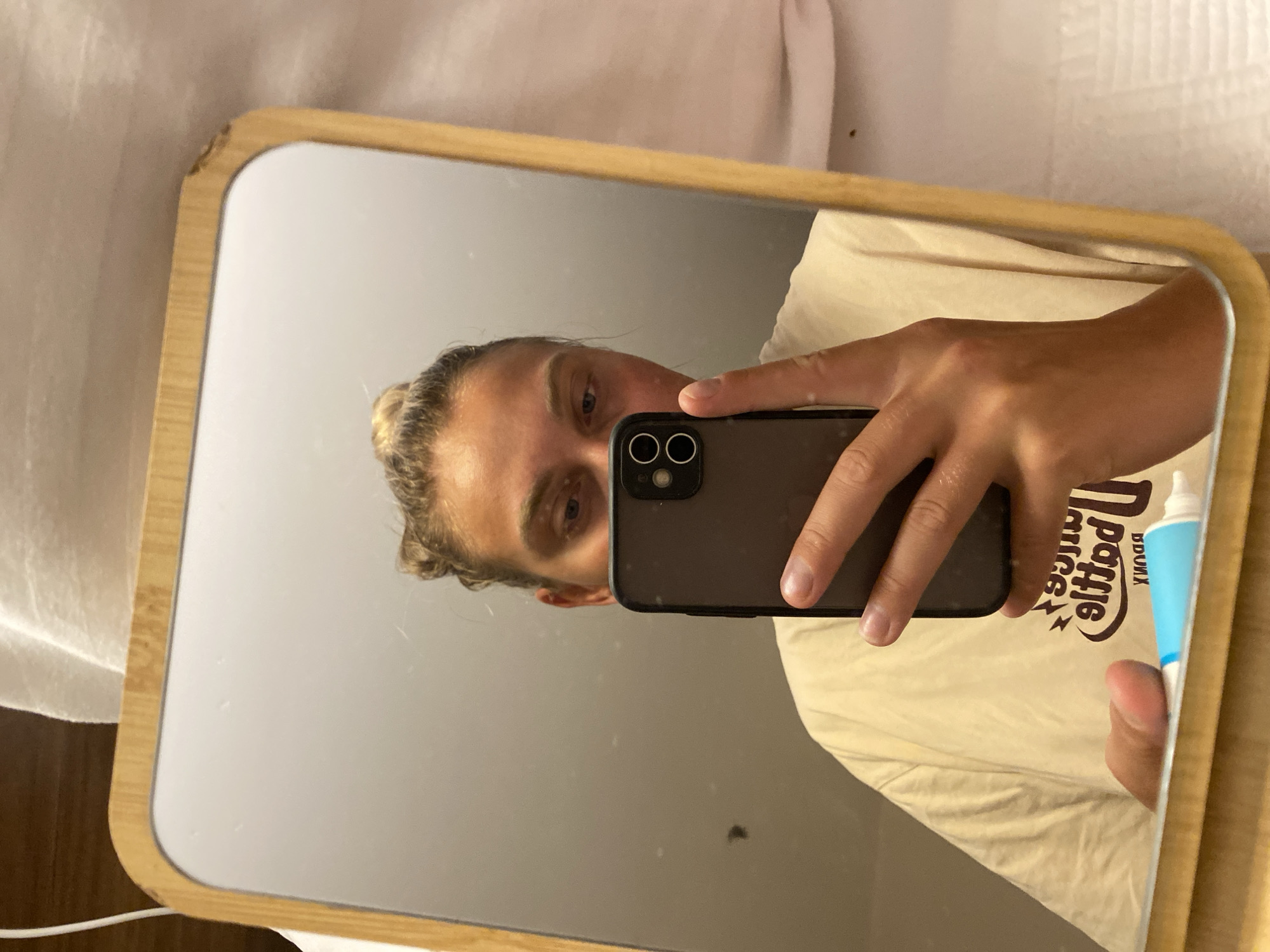 A person is taking a selfie in a mirror. The person is holding a smartphone in their right hand and a tube of toothpaste in their left hand. The person is wearing a light-colored t-shirt. The mirror has a wooden frame. The background appears to be a bathroom. There are no indications of the location in Aksu, Turkey.