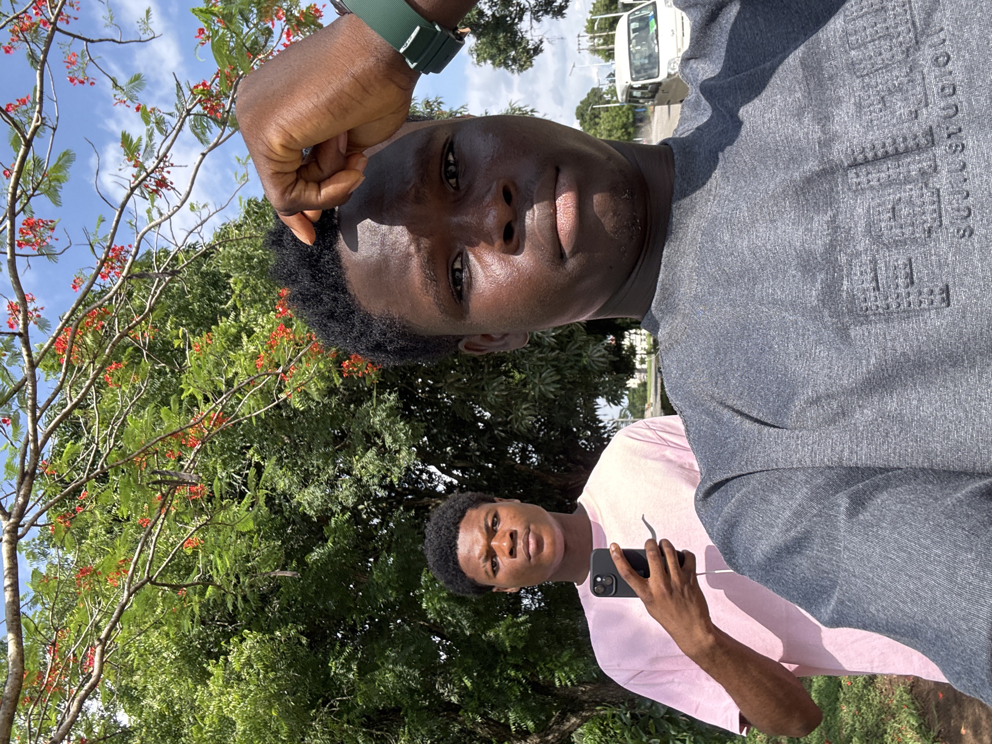 Two young men are depicted outdoors in bright daylight in Ihiagwa, Nigeria. The primary subject, positioned closer to the viewer, is taking a selfie. He has dark skin, short dark curly hair, and is wearing a grey t-shirt with a green wristband on his left arm. His t-shirt features embossed text, partially readable, which includes "SULSTUDIOS" and likely other words like "FUTURE" or "ESSENTIALS".
Behind him and slightly to his right stands a second young man, also with dark skin and short dark curly hair. He is wearing a light pink t-shirt and holds a black smartphone with a dual-camera system in his right hand. Both individuals have neutral expressions.
The setting is vibrant with lush green trees and foliage, some bearing small red flowers, especially prominent on the left side of the frame. Above the greenery, a clear blue sky with sparse white clouds is visible. In the upper background, a white vehicle, resembling a bus or van, is present on a paved surface, suggesting a road or parking area. The strong sunlight indicates it is daytime, likely midday or early afternoon.