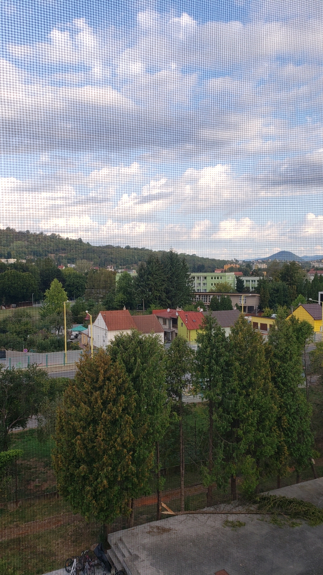The image depicts a view from a window with a screen, looking out over a residential area in Prešov, Slovakia. In the foreground, there are several large evergreen trees and a concrete structure. A cluster of bicycles is visible on the concrete. Beyond the trees, there are houses with red and yellow roofs, and further in the background, a light green apartment building stands out. The landscape is hilly, covered with trees, and a distant mountain peak is visible on the horizon. The sky is filled with scattered clouds.