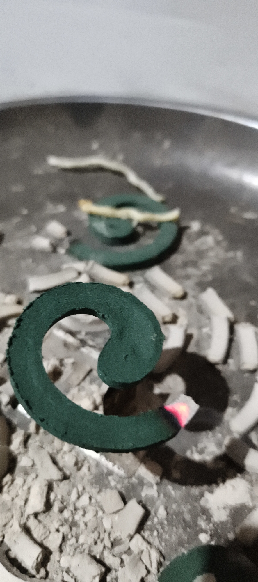 In Muaro Jambi, Indonesia, a close-up shot focuses on two partially burned mosquito repellent coils placed on a metallic surface covered in ash and remnants of extinguished coils. One coil, prominently in the foreground, is dark green and exhibits a glowing red ember at its tip, indicating it is actively burning. The other coil, further back, is also green but appears fully extinguished. Both coils are arranged in a spiral pattern, a characteristic shape for this type of pest control product. The surrounding ash and fragments suggest a history of use, with pieces of broken coils scattered around the main subjects. The lighting is dim, casting shadows and highlighting the glowing ember.