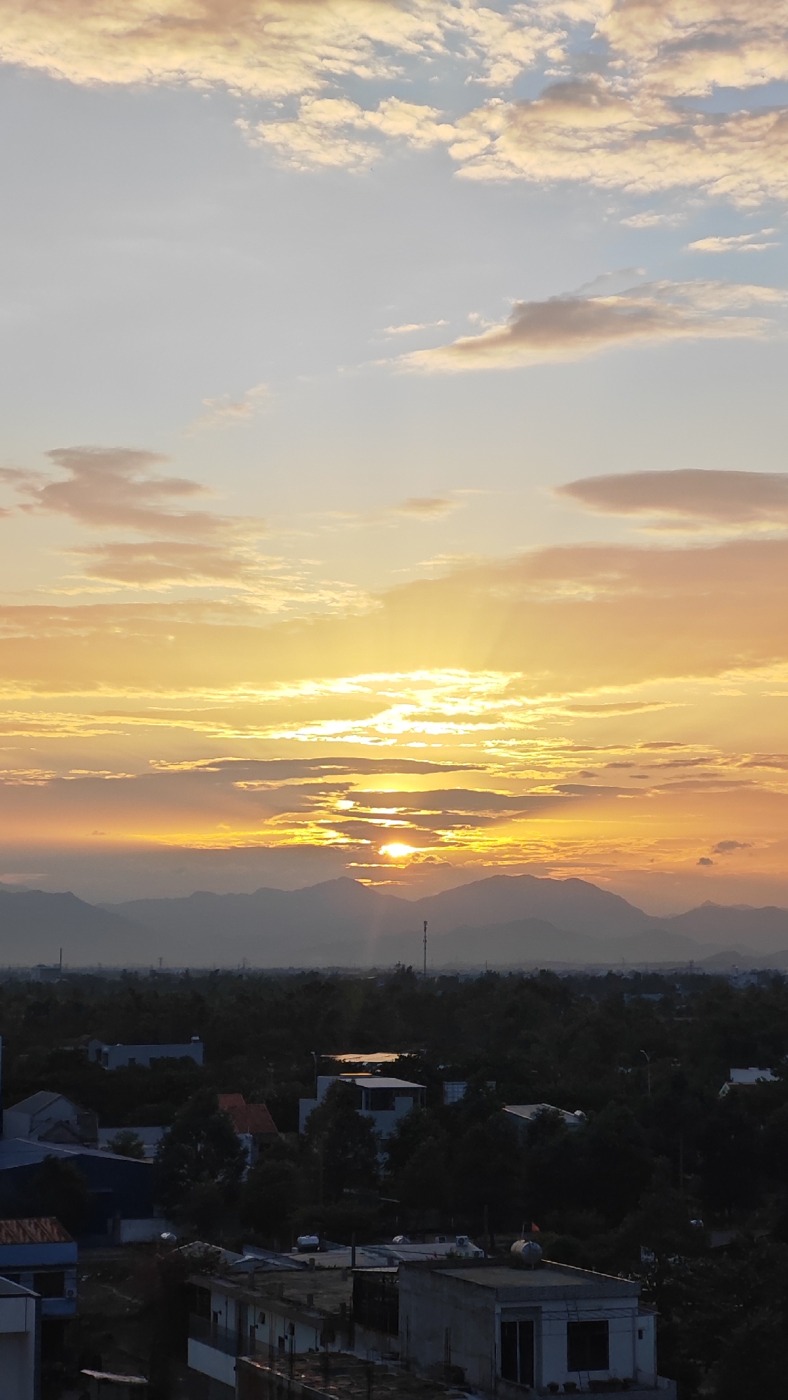 The image captures a sunset over a suburban area of Da Nang, Vietnam. The sky is dominated by warm hues of orange and yellow, with streaks of sunlight breaking through scattered clouds. In the distance, a range of mountains is silhouetted against the vibrant sky. Below, the landscape transitions into a dense expanse of dark green trees, interspersed with the roofs of residential buildings. The buildings are of various sizes and designs, some appearing modern with white facades, while others have more traditional tiled roofs. The scene is tranquil, with no visible people or active elements, suggesting a quiet moment at dusk. The overall impression is one of peaceful natural beauty meeting urban development.