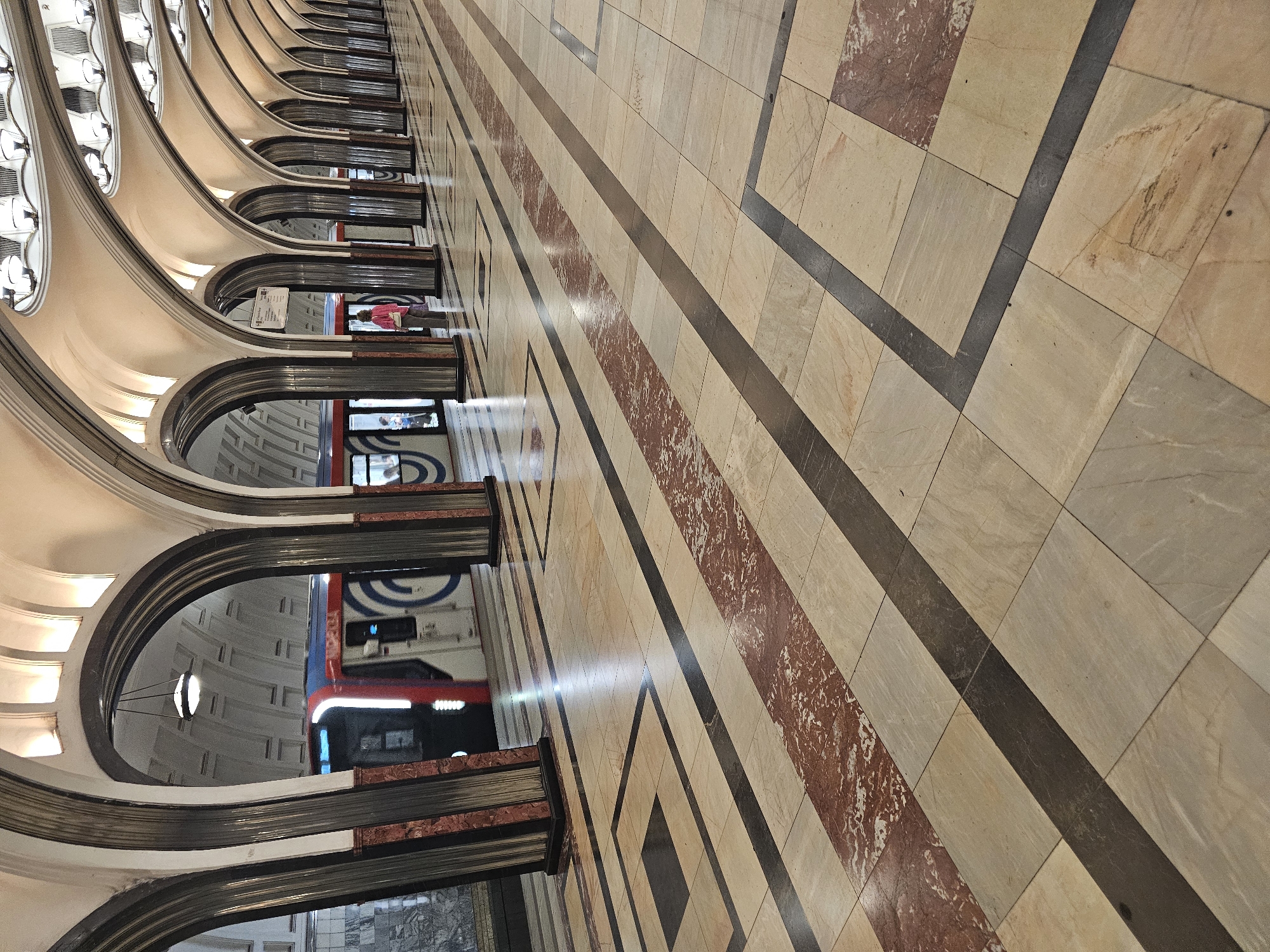 This image depicts the interior of a Moscow Metro station, likely the Mayakovskaya station, characterized by its opulent Art Deco architecture. The scene captures a long, arched hall with polished marble floors. Several imposing, fluted columns with dark grey and red marble detailing line the platform, supporting a series of grand, illuminated arches that form the ceiling.
In the background, a train with red and white carriages is visible, stopped at the platform. The doors are open, and a few passengers can be seen. One individual, wearing a bright pink jacket, stands on the platform looking towards the train. The floor is paved with large square tiles of varying neutral tones, accented by dark grey and reddish marble strips arranged in geometric patterns, creating a sense of depth and perspective. The overall atmosphere is one of grandeur and historical significance, typical of Moscow's ornate metro system.