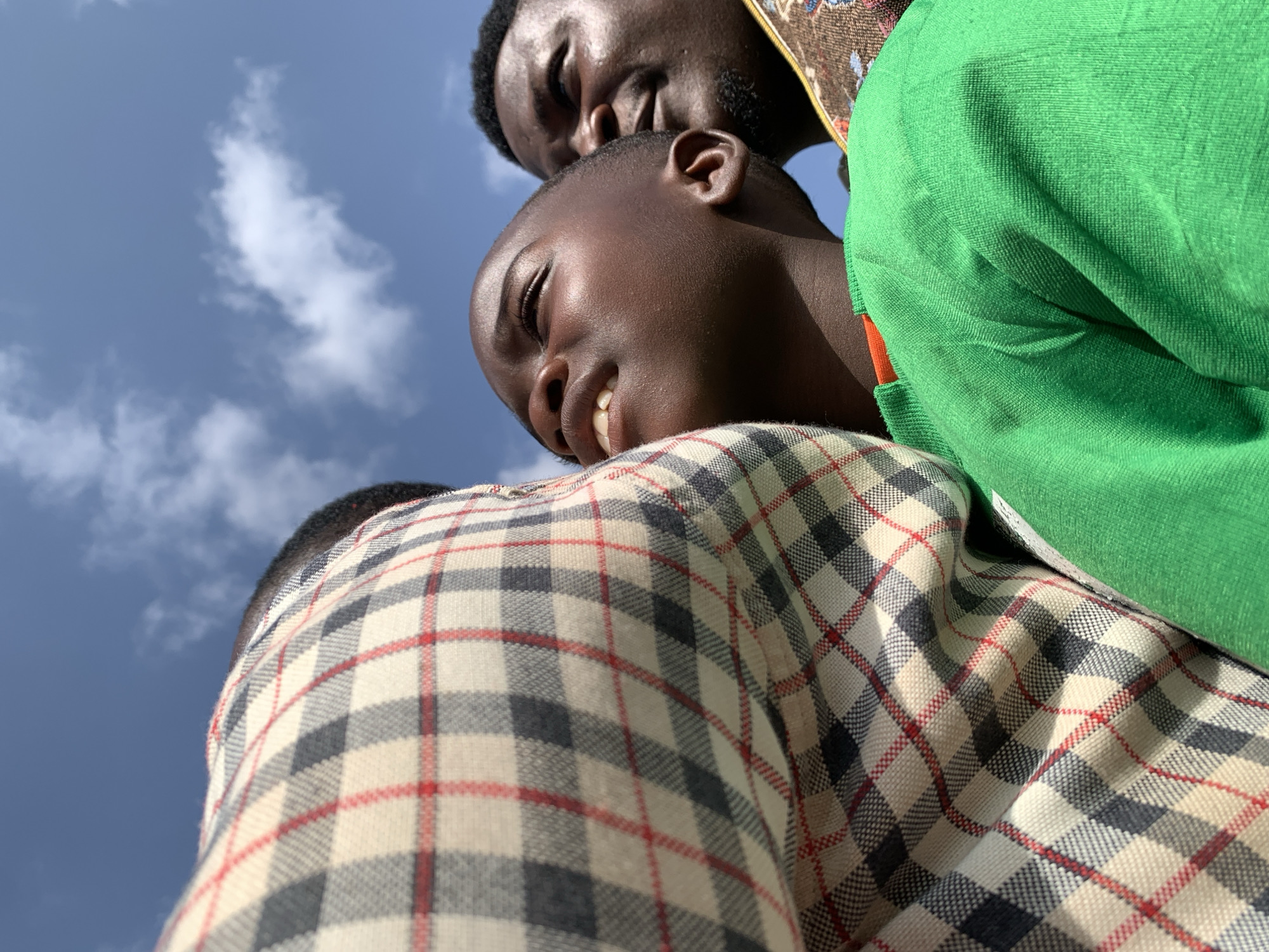 Three people are in the image, taken from a low angle looking up at them against a blue sky with scattered white clouds. The person closest to the camera wears a plaid shirt with a white background and red and gray lines. Above this person, another person is looking down, with a partially closed eye and a slight smile. A third person is partially visible to the right, wearing a green top. It is daytime and the location is presumed to be in Akwanga, Nigeria.