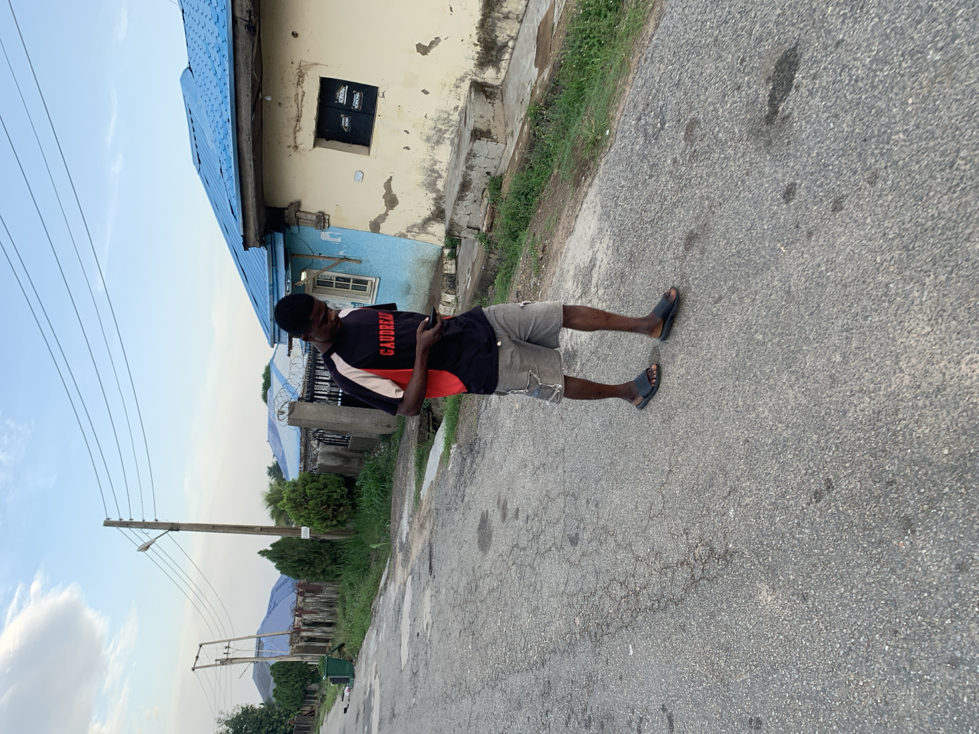 A man stands on an asphalt road, facing slightly away from the camera, holding a dark rectangular object (likely a phone). He is wearing a black t-shirt with red and white accents and the word "GAUDREAU" visible, along with grey shorts and dark-colored sandals. A building with a blue roof and partially deteriorated beige walls is in the background. There's a window with a security grille on the upper part of the building. A power pole with multiple wires is visible against a cloudy sky. A blue roof is on a house behind a barbed wire fence. The scene is most likely in a residential area. A distant green hilly landscape is visible. The asphalt road shows signs of wear and tear. Given the presence of the name "GAUDREAU" on the shirt and a common street name, it is most likely not in the city of Abuja, Nigeria.