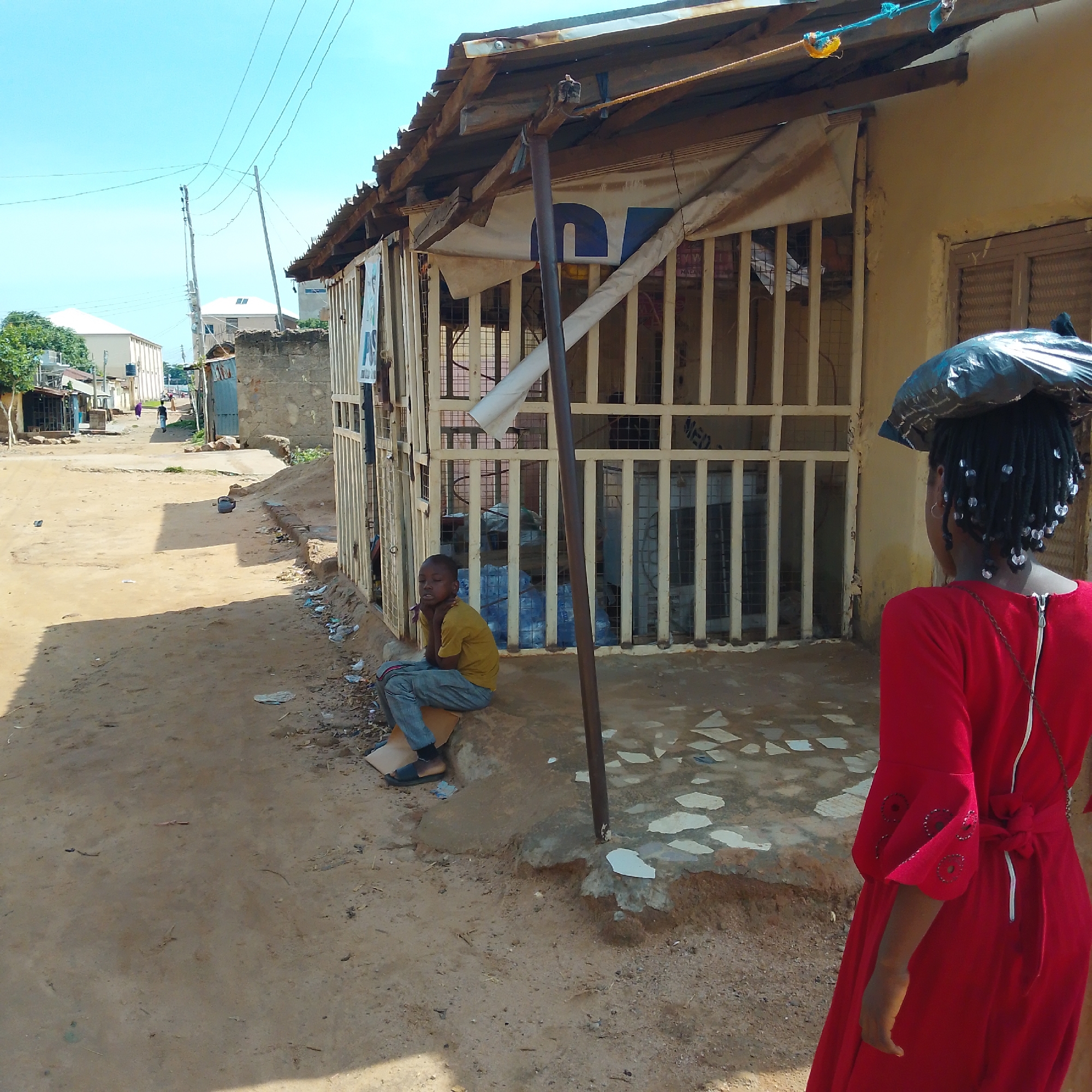 In Pantame, Nigeria, on a dusty, sunlit street, a young boy in a yellow t-shirt and gray pants sits on a concrete ledge outside a shop. The shop has a corrugated metal roof and bars on its windows. In the foreground, a person wearing a bright red dress walks away from the camera, carrying a large black plastic bag on their head. The street is unpaved, with scattered debris and a few scattered plastic bottles visible on the ground. In the background, more buildings and a few people can be seen, along with power lines stretching across the clear blue sky.