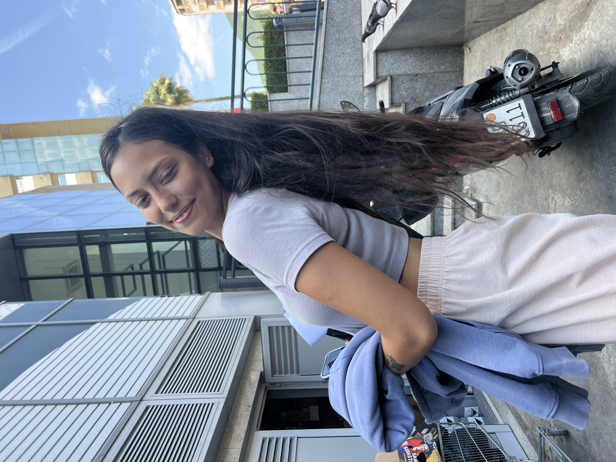 A young woman with long, dark hair is standing outside a modern building in Budva, Montenegro. She is wearing a light gray crop top and light beige sweatpants, holding a blue sweatshirt in her arms. Her eyes are closed, and she has a slight smile on her face, leaning her head back. In the background, the building features large glass windows and horizontal silver siding. To her right, a black scooter with a license plate is parked. Pigeons are visible on a concrete ledge near the scooter. The sky is blue with scattered clouds, and a palm tree is present in the distance.