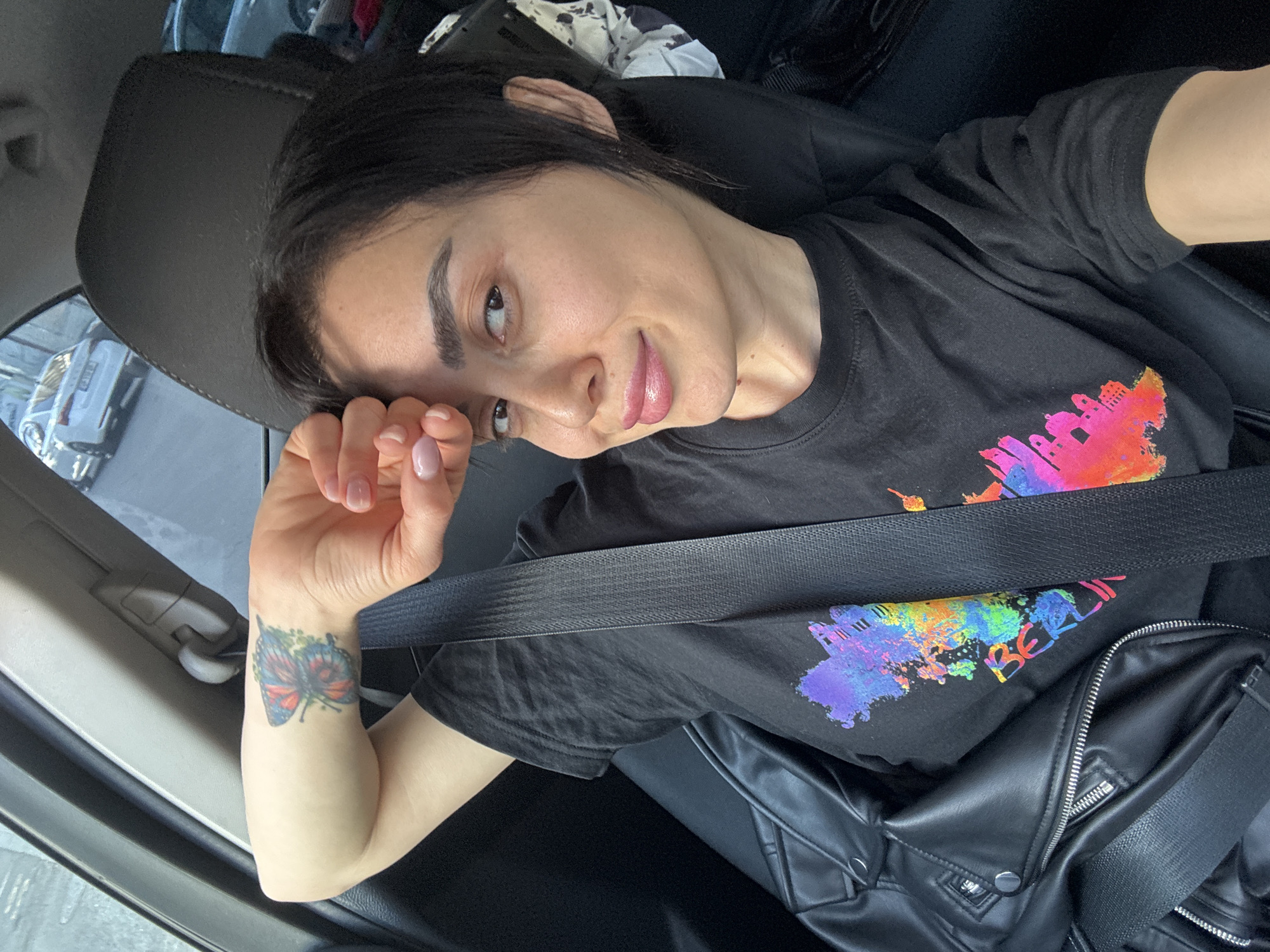 A woman is sitting in the driver's seat of a car, wearing a black t-shirt with a colorful graphic that says "Berlin". She is wearing a seatbelt and a black leather jacket is lying next to her. The woman has short dark hair and light skin. She has a butterfly tattoo on her left wrist. The car is parked on a street with other cars visible outside the window. The exact location in Tbilisi, Georgia cannot be determined from the image.