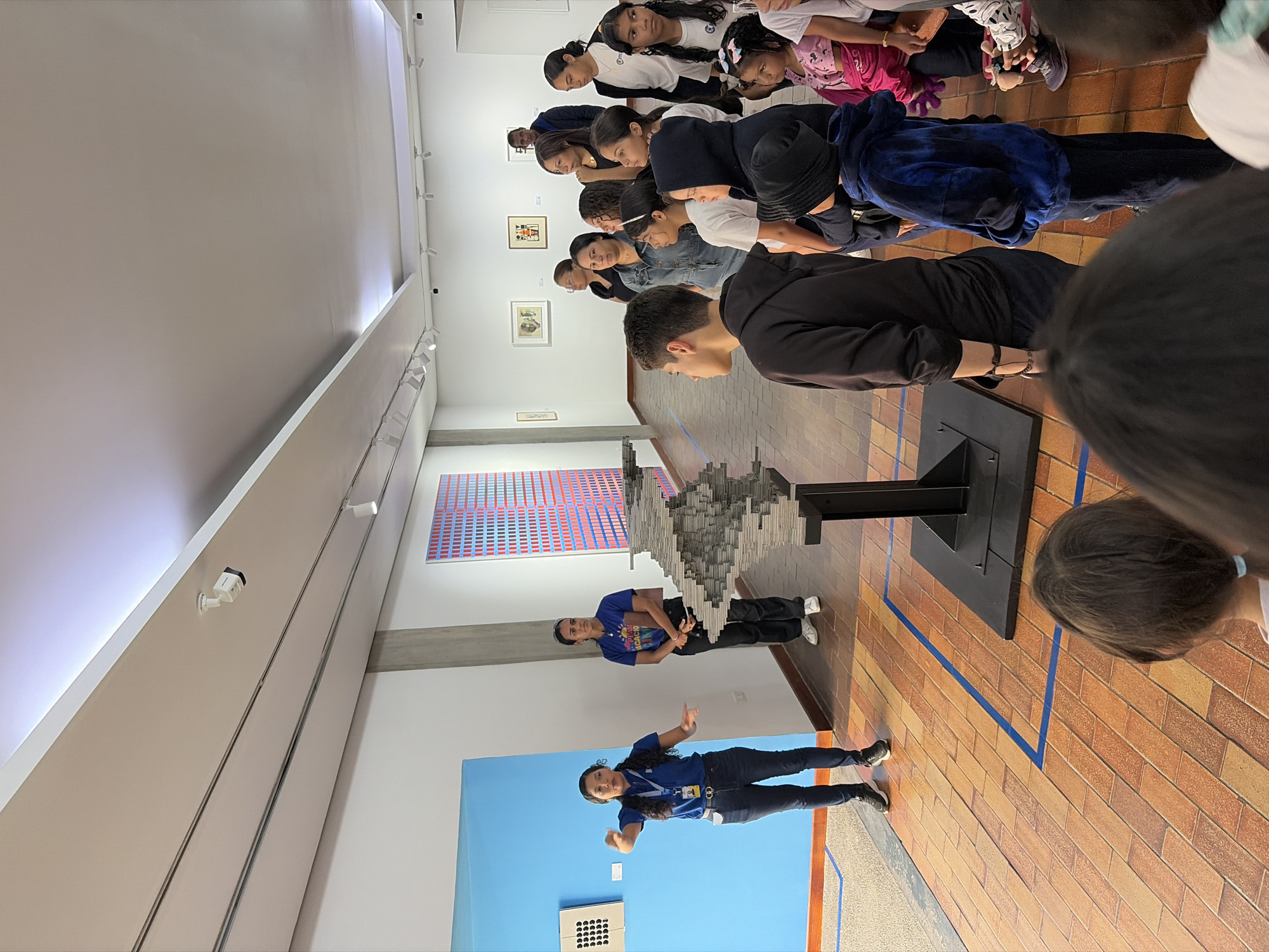 A group of approximately 12 individuals, including children and adults, are gathered in an art gallery. The individuals are arranged along a blue line on the floor, looking toward a sculpture positioned in the center of the room, they all seem to be observing it. One adult is standing near the sculpture, giving information about it. Another adult stands to the left, wearing a blue shirt. The walls are white. There are several pieces of artwork, including one that has red and blue squares. The floor is brick. The location is unknown, not Ciudad Bolivar, Venezuela.