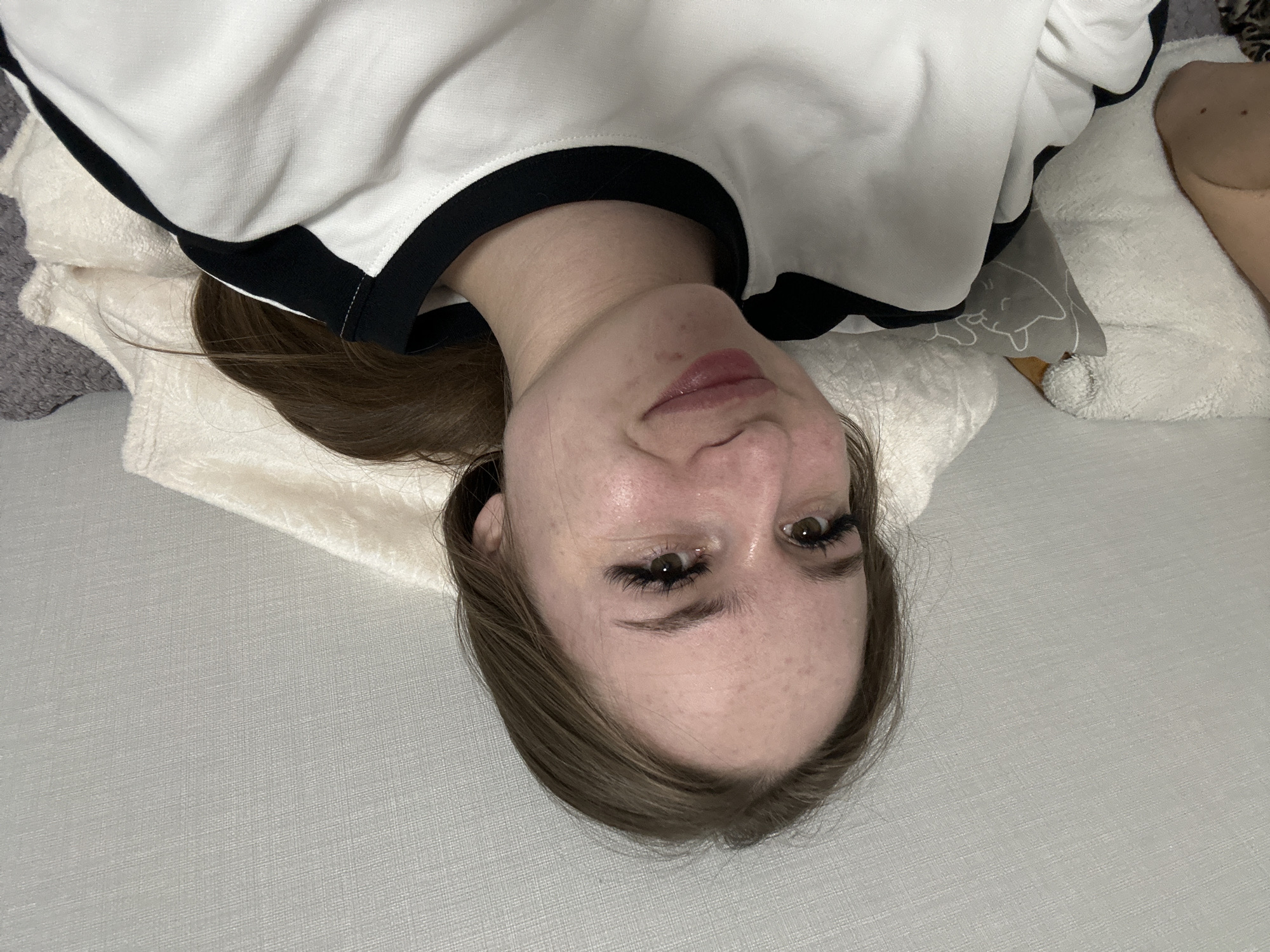 A young person, likely female, is depicted lying down in an indoor setting, with her head inverted in the frame. She looks directly at the viewer with a neutral to slightly smiling expression. Her light brown hair spreads around her head. She has fair skin with visible freckles and dark eyes, complemented by prominent, long eyelashes and defined eyebrows. Her lips are slightly parted and appear glossed.
She is wearing a casual white top with black trim around the neck and shoulders. Her head rests on a white, fluffy pillow. Adjacent to her, a grey pillow with a white cartoon cat design is partially visible. To the right of the frame, a bare arm or shoulder rests on a soft, light-colored surface, possibly another pillow or blanket. The main surface she is lying on is a light-colored, textured fabric, resembling a bed sheet or sofa covering. The scene suggests a relaxed atmosphere, likely in a bedroom or living room. No text is visible in the image.