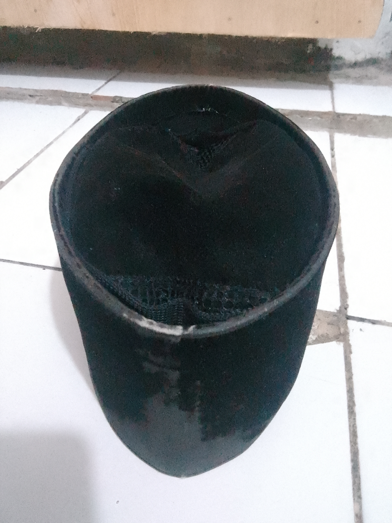 A black, kufi-style hat is positioned on a tiled floor. The hat is cylindrical with a flat top, and the material appears to be fabric. The top of the hat has some stitching details creating a pattern. The hat is the sole object in the foreground, and its placement suggests it has been set down. The background shows a floor composed of white square tiles, with grout lines visible. Above the tiled floor, there's a wooden surface, possibly a shelf or part of a structure, which is lighter in color. The lighting suggests an indoor setting, possibly with natural light filtering in. The scene is still and lacks any human presence or apparent activity, focusing solely on the presentation of the hat. There is no visible text in the image. The location context is Cisarua, Indonesia.