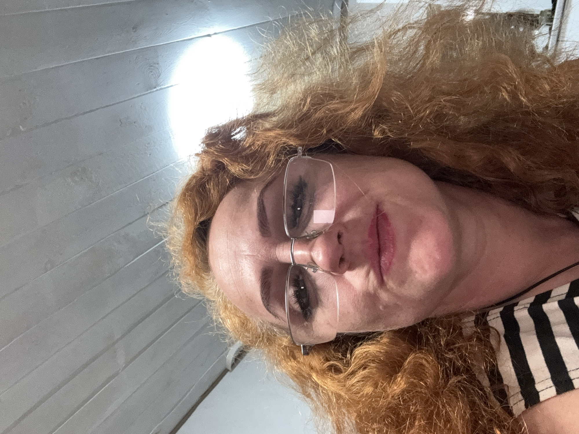 The image shows a woman with red, curly hair wearing eyeglasses. She is positioned at an unusual angle, suggesting the photo was taken from above. Part of a white, paneled wall is visible in the upper left corner. The woman is wearing a black and white striped top. The background and positioning give a slightly disorienting effect. The location in the city of Chaikovsky, Russia is not evident from the visual information available in the image.